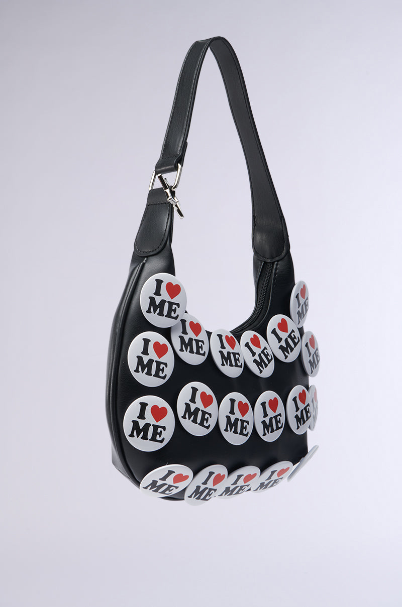 The I REALLY DO PIN PURSE from the AKIRA Label is a chic black faux leather accessory featuring "I ♥ ME" round buttons. Its curved design, single handle, and minimalist style stand out against a soft gradient background.