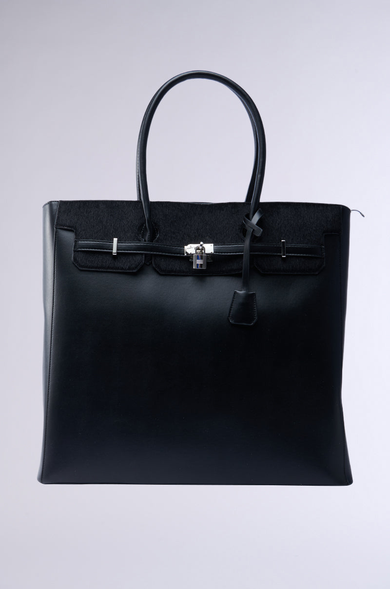 The FEEL ME OVERSIZED TOTE is a sleek black faux leather handbag with two handles and a small metal lock. It offers a spacious, structured rectangular interior with subtle stitching details set against a soft gradient background.