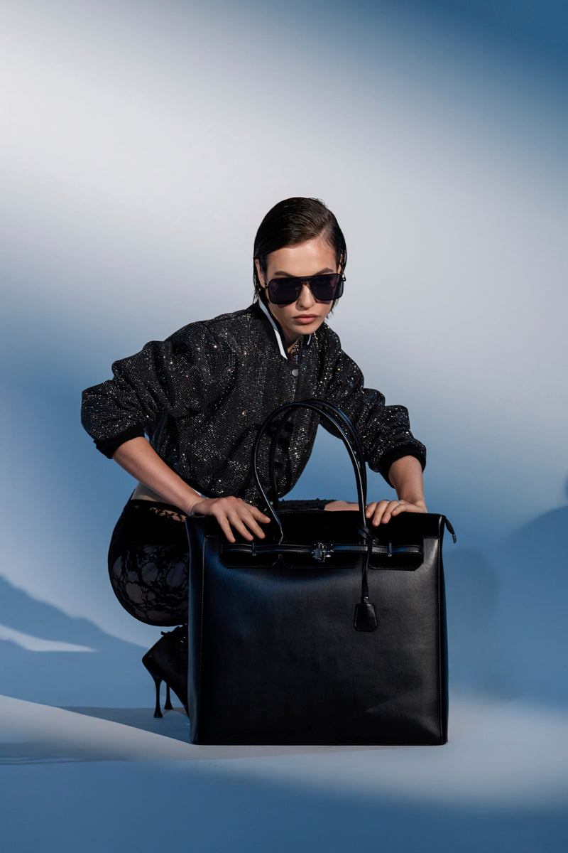 A stylish woman in a sparkly black jacket, sunglasses, and high heels poses with the FEEL ME OVERSIZED TOTE, a spacious faux leather handbag, against a gradient blue background.