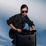 A stylish woman in a sparkly black jacket, sunglasses, and high heels poses with the FEEL ME OVERSIZED TOTE, a spacious faux leather handbag, against a gradient blue background.
