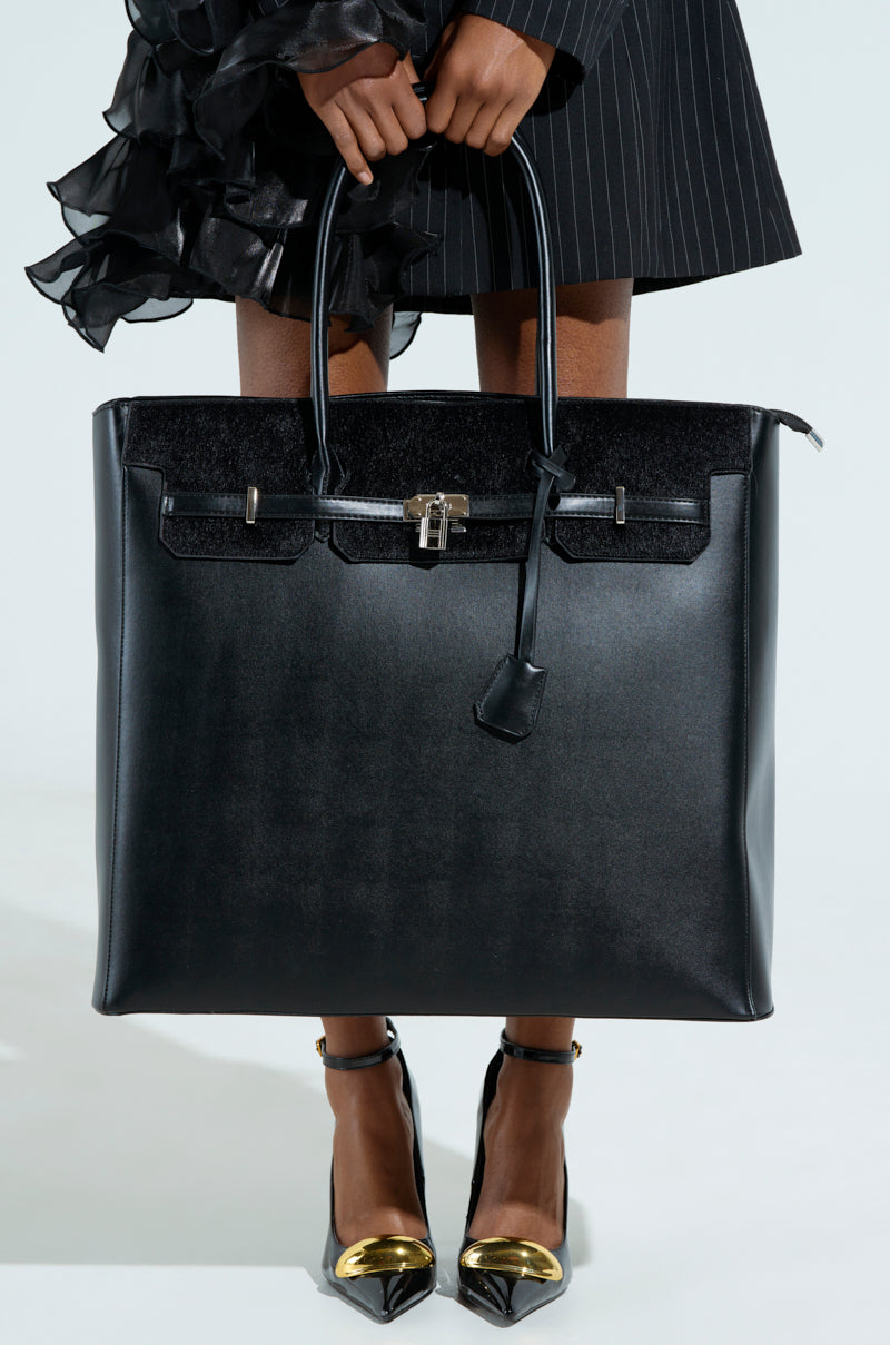 In a pinstripe outfit, a person carries the FEEL ME OVERSIZED TOTE crafted from faux leather with a metal lock, paired with glossy black heels featuring golden tips against a plain white background.
