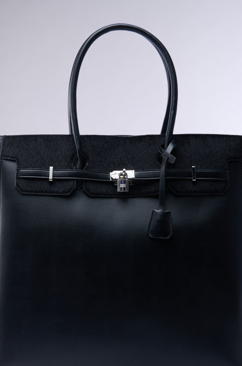 The FEEL ME OVERSIZED TOTE is a sleek black faux leather handbag with a structured design, two handles, and a silver clasp lock. It features a subtle textured finish and spacious interior, offering both elegance and functionality against a simple gradient background.
