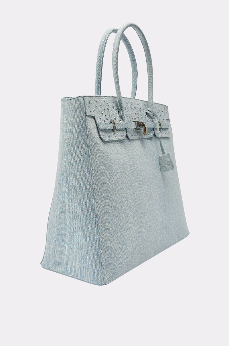 The DENIM AND DIAMONDS OVERSIZED TOTE is a light blue textured handbag with two handles, a structured rectangular shape, subtle rhinestone accents, a front flap, and silver-tone hardware, displayed angled on a plain white background. 4