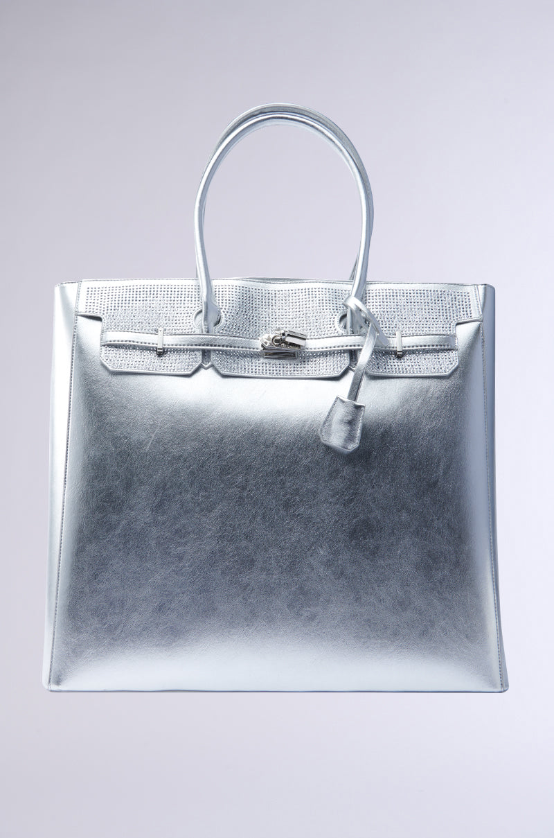 The GET IN MY BAG OVERSIZED TOTE is a silver designer handbag made from faux patent leather, featuring dual handles, a textured flap, and a front lock accent. Its elegant, shiny surface beautifully reflects light, enhancing its luxury appeal against a plain backdrop. 1