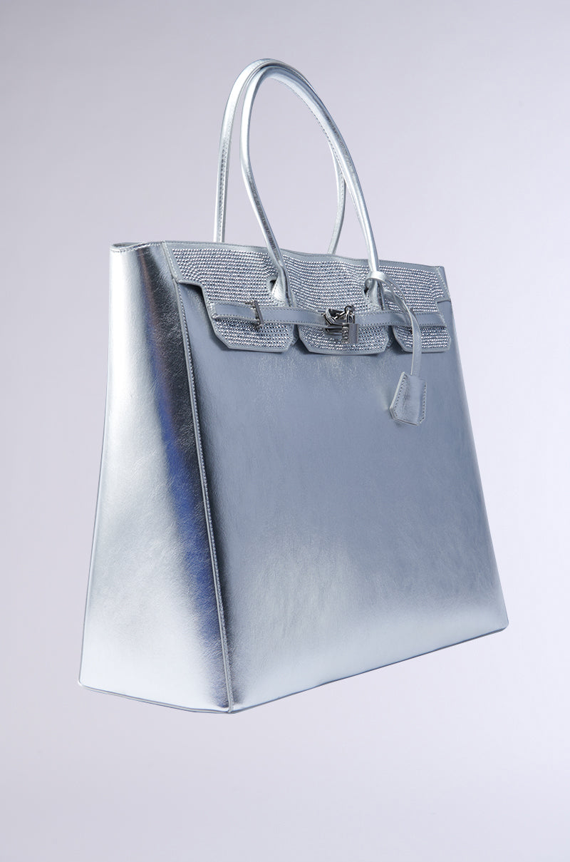 The GET IN MY BAG OVERSIZED TOTE is a silver faux patent leather handbag featuring a structured, shiny design with dual handles, a flap closure with a unique clasp, and a small rhinestone pendant. The bag is showcased against a gradient background with soft lighting. 3