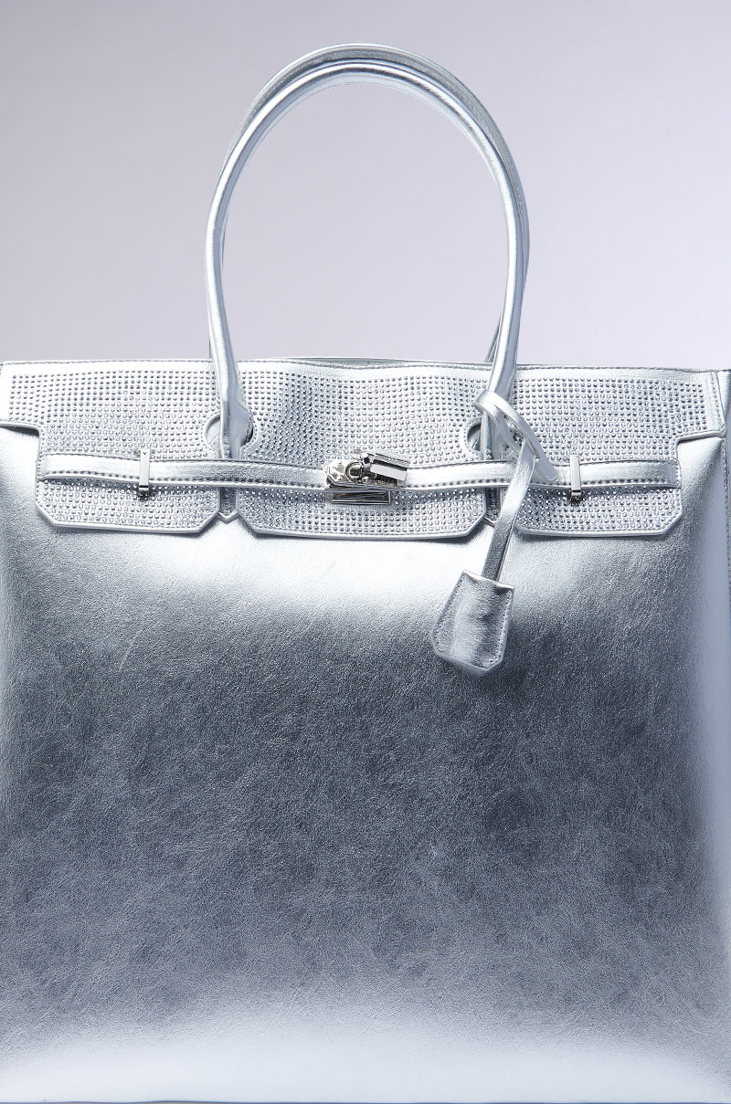 The GET IN MY BAG OVERSIZED TOTE is a sleek, silver faux patent leather handbag with a textured finish, two handles, a padlock with a leather tag, and a top zipper detail, set against a plain light gradient background. 4