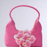 The IN BLOOM PINK PURSE is a pink floral handbag with simple handles, adorned with a cluster of pink and white orchids on its plain faux leather background, highlighting its elegant floral design.