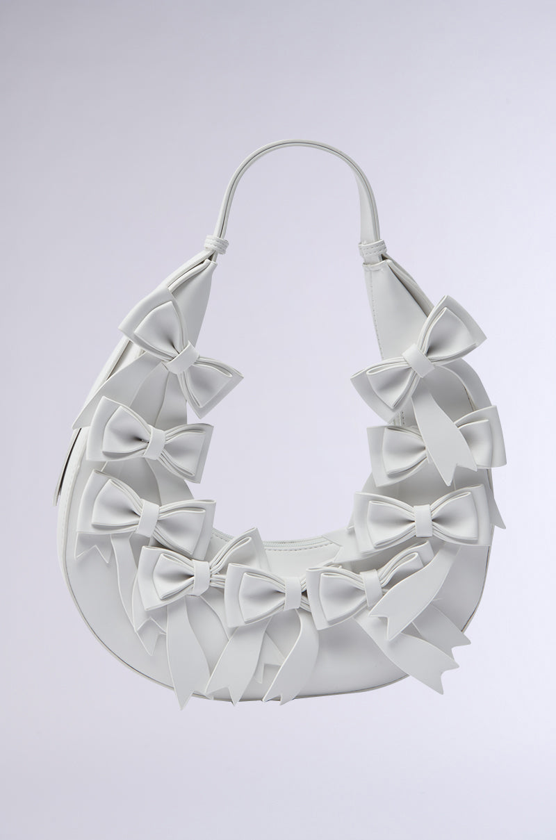 The TIE IT WITH A BOW PURSE is a white faux leather crescent-shaped handbag adorned with multiple white bows in varying sizes on the front and handle, displayed against a light gray background.