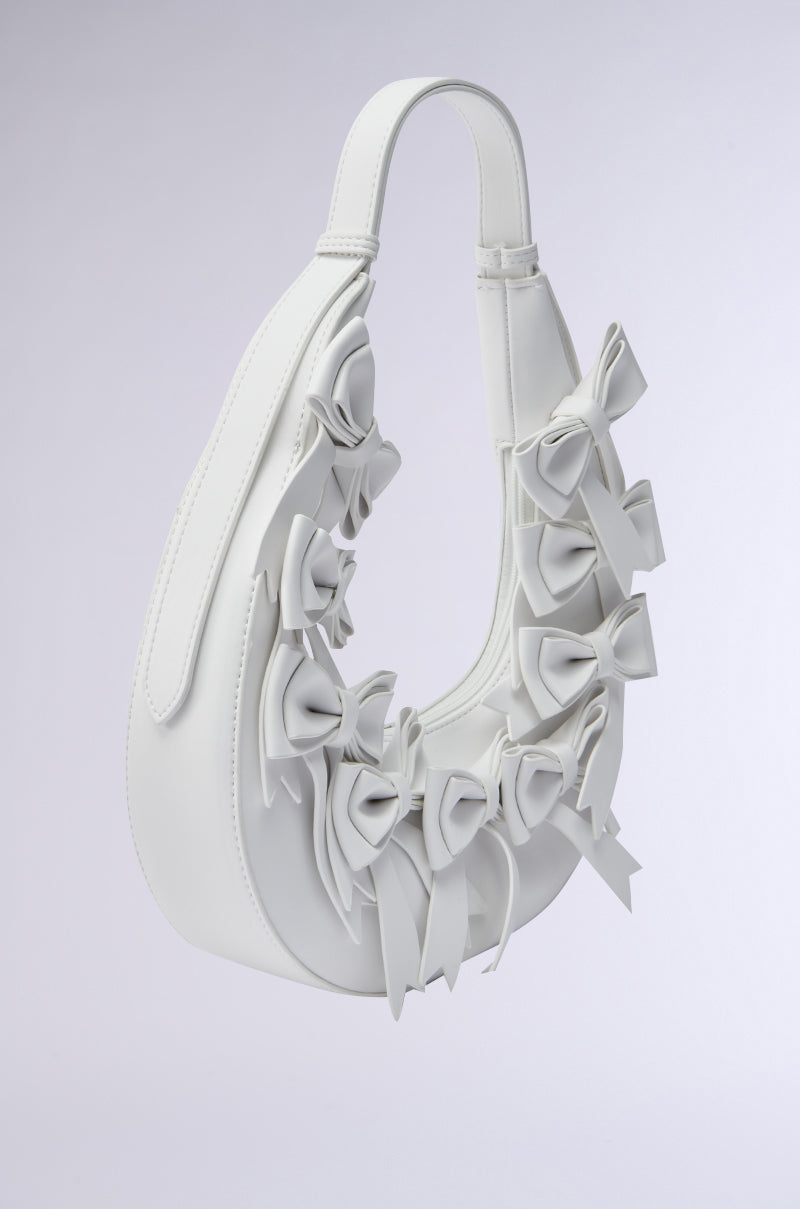 The TIE IT WITH A BOW PURSE is a white crescent-shaped handbag in faux leather with a curved handle and several bows on the front, shown against a light gray background.