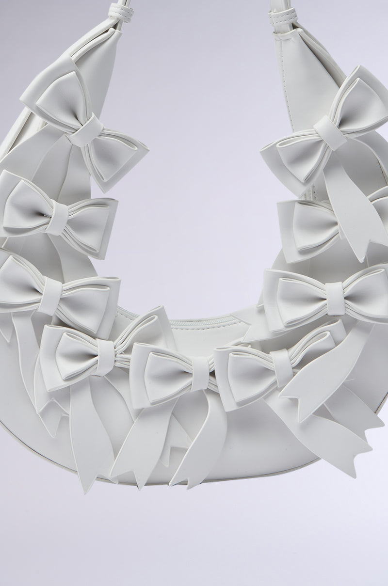 The TIE IT WITH A BOW PURSE is a white, crescent-shaped faux leather handbag featuring multiple white bows on the front, displayed against a simple light background.