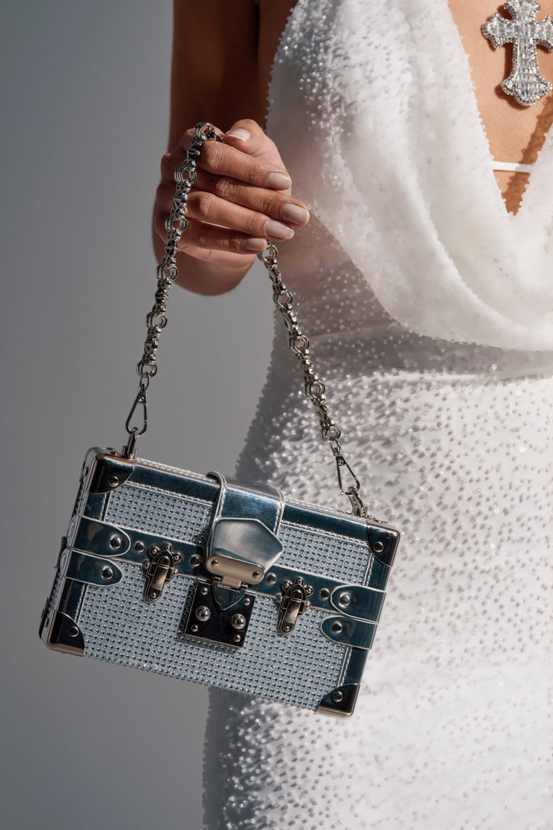 A person in a white beaded dress and cross necklace holds the LOU BLING TRUNK BAG, adorned with silver rhinestones, metal clasps, and a chic chain handle.