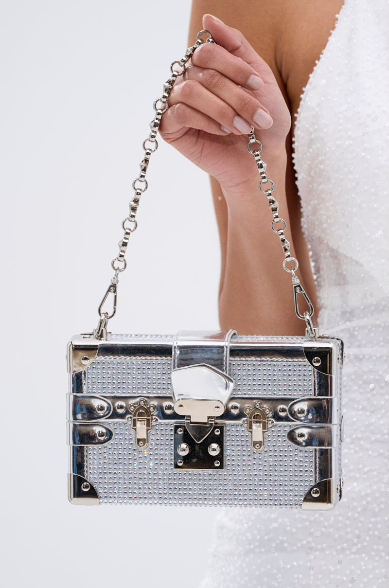 A person in a white textured dress holds the LOU BLING TRUNK BAG—a small, box-shaped statement purse covered in silver rhinestones with a chain handle and decorative clasps.