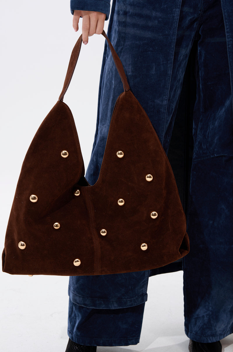 A person in dark blue velvet pants holds the TAYLOR SUEDE BALL TOTE, an oversized brown suede bag with shiny gold studs, two handles, and an angular silhouette, standing out against a plain white background.