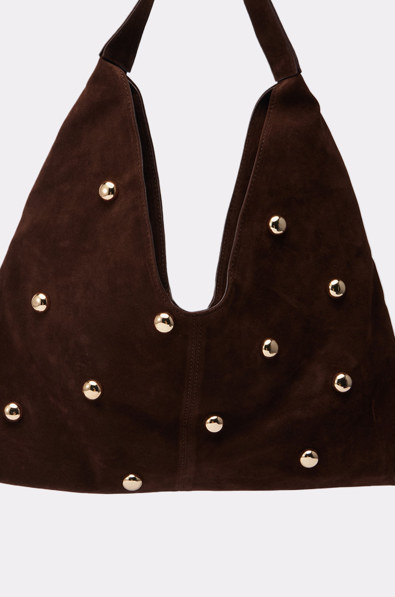The TAYLOR SUEDE BALL TOTE is a dark brown faux suede oversized hobo bag featuring a wide strap and evenly spaced gold round studs, shown against a plain light background.