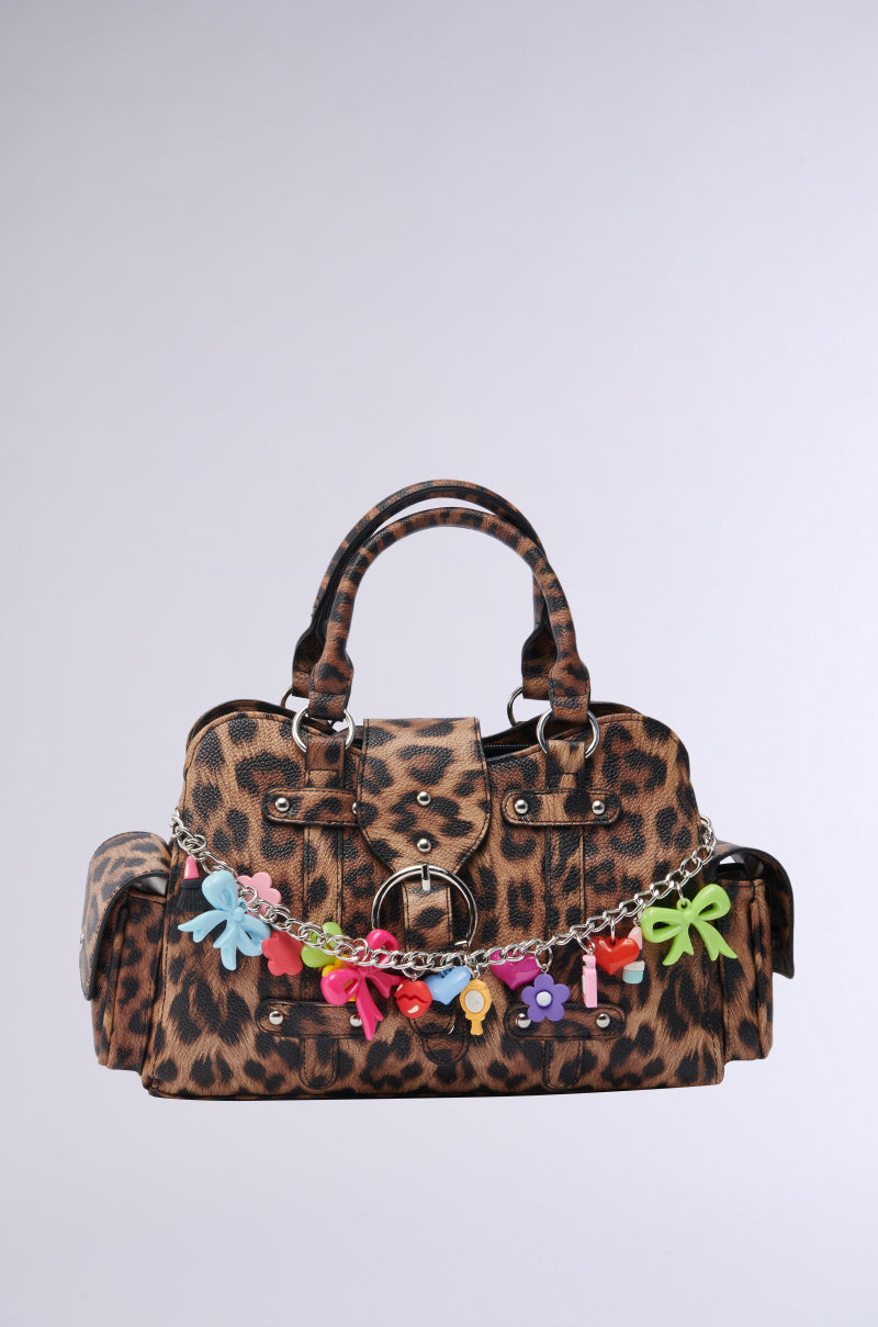 The ITS BETTER WILD PURSE features a cheetah print, double handles, side pockets, and a silver chain decorated with Y2K-style flower, bow, and heart charms, all set against a plain light background. 1