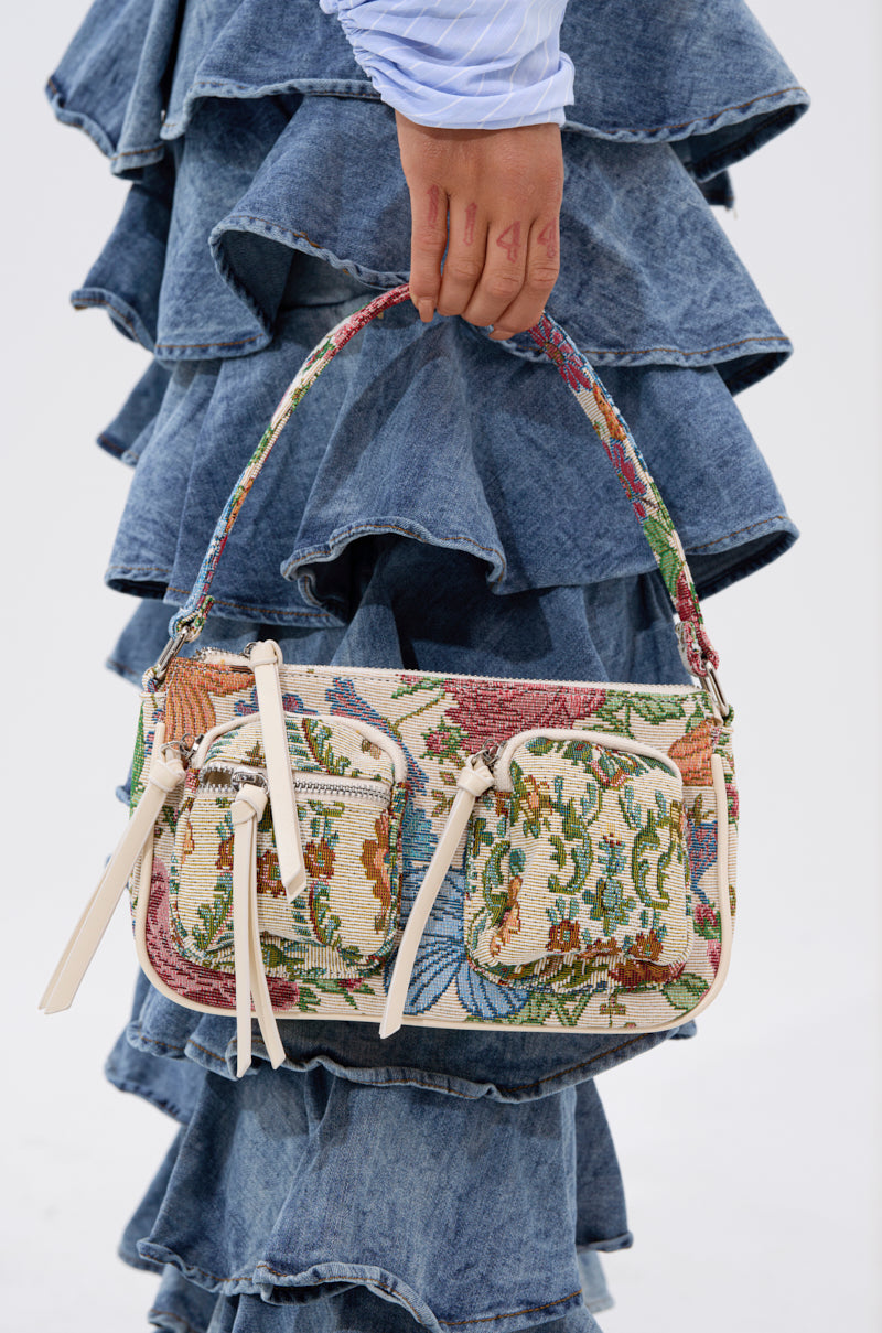 A person holds THE REAL KNOW PURSE, a small floral tapestry bag with three zippered pockets, ornate patterns, and cargo-style details, against a plain background while wearing a ruffled denim skirt. 1