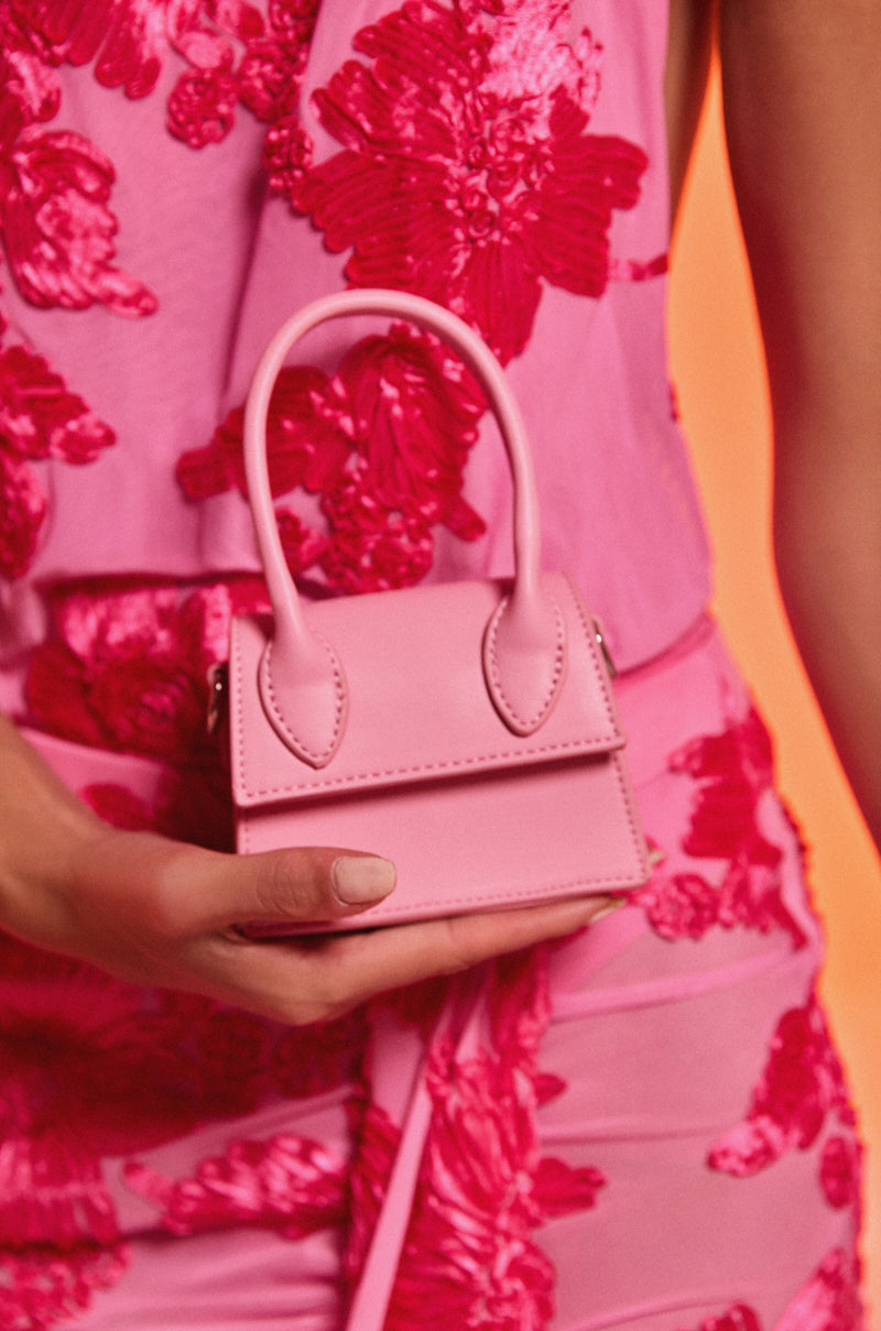 A person in a pink dress with red floral patterns holds the JACKIE PURSE, a small faux leather bag with two handles, against a peach-colored background.