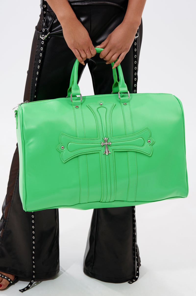 A person in black lace-up leather pants holds the SHE DIFFERENT DUFFEL BAG by AKIRA, a bold lime green faux leather duffel featuring a decorative cross design and metal embellishments on the front.