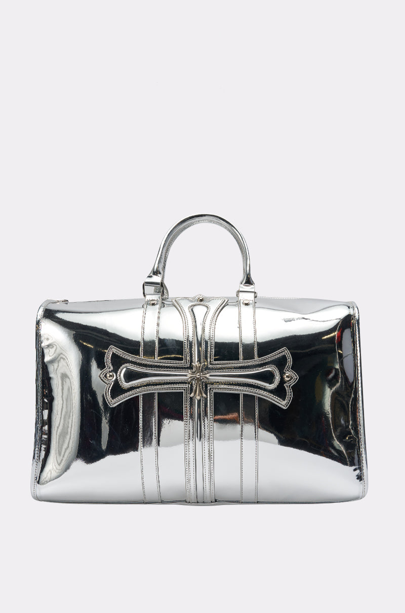 The IM CHROME DUFFEL is an oversized, shiny silver metallic bag with dual handles and a cross-shaped appliqué on the front, displayed on a plain white background.