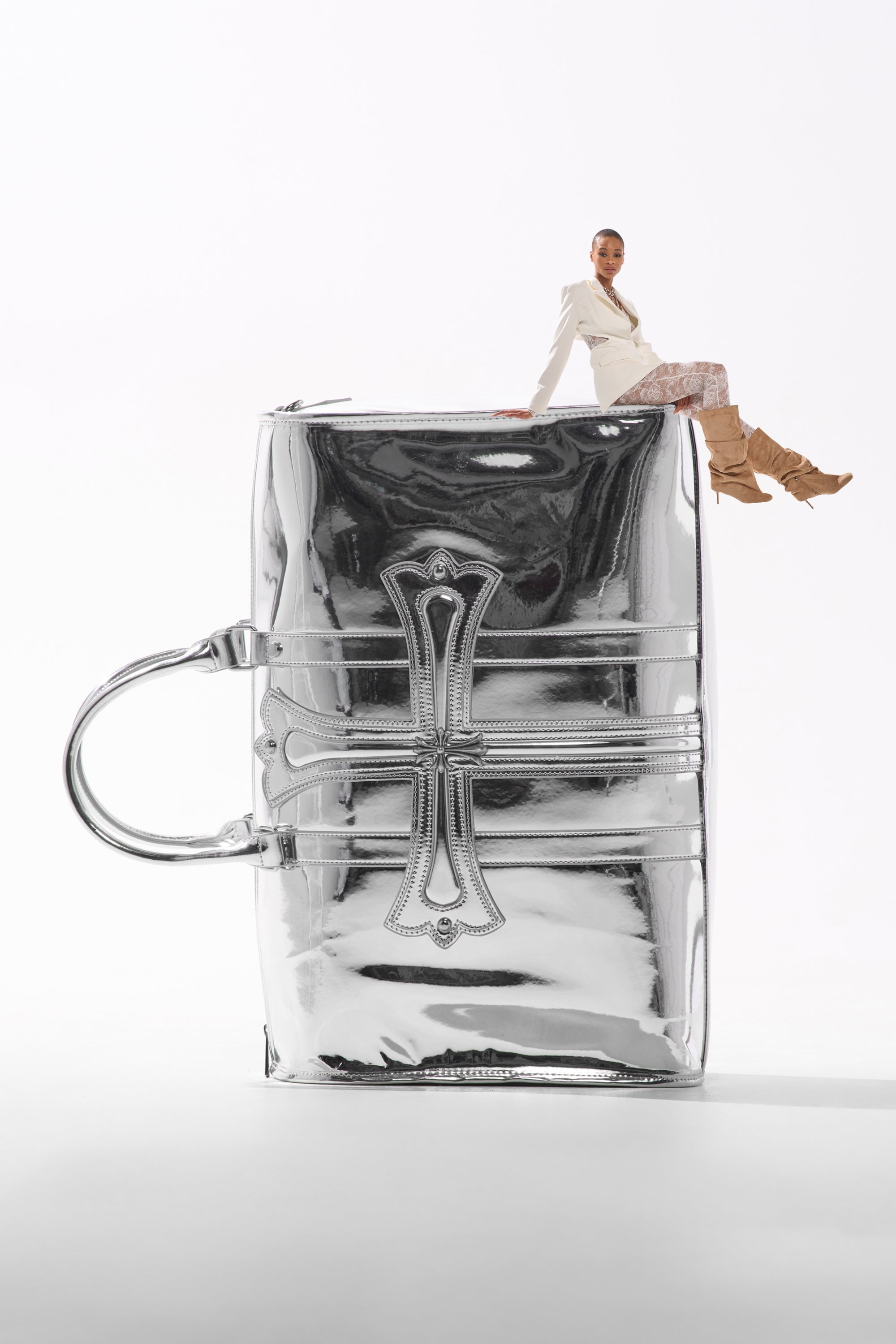 A woman in stylish clothing sits atop the IM CHROME DUFFEL, a large silver metallic bag with intricate detailing, creating a playful and surreal scene against a white background.