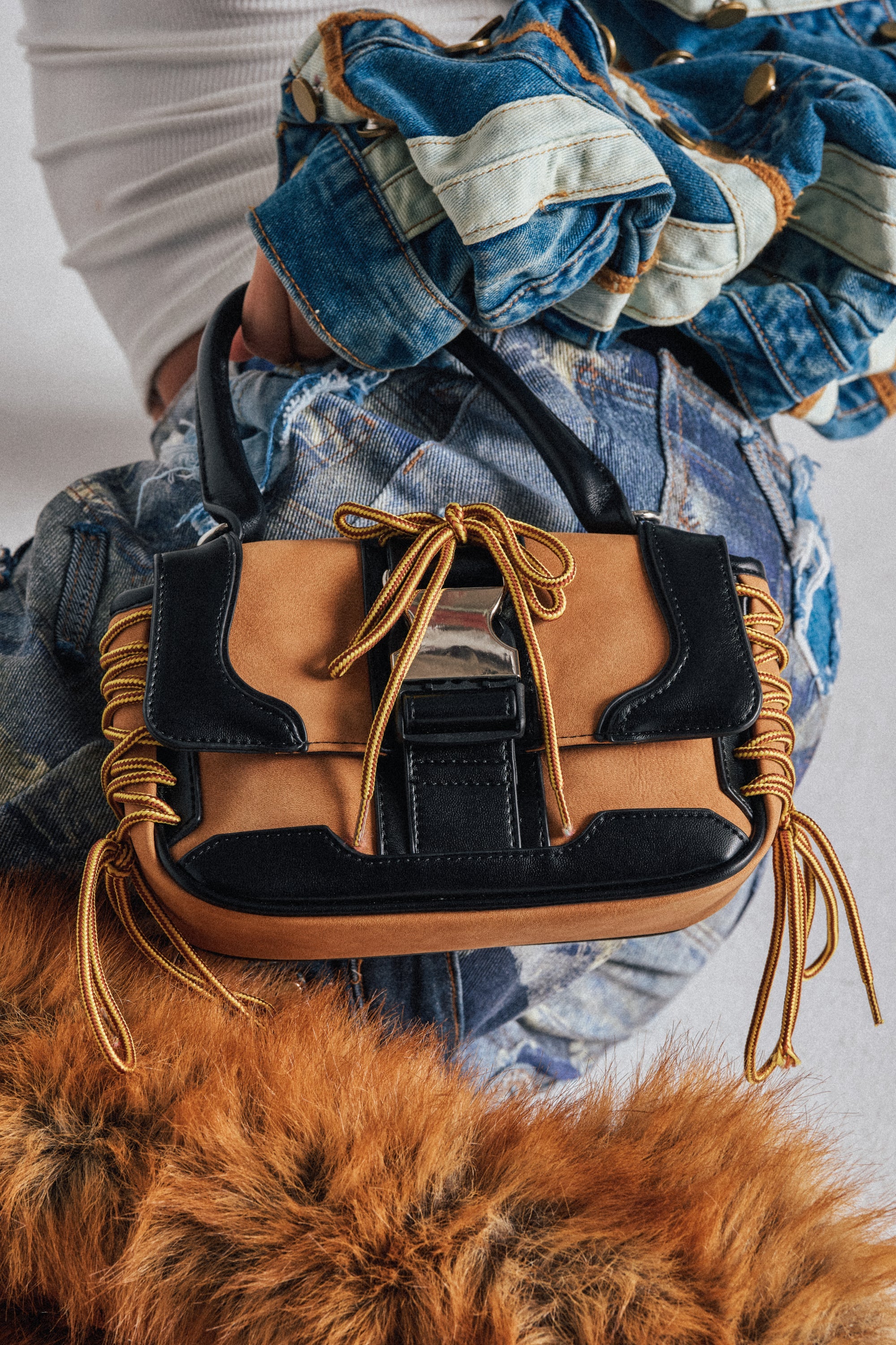 The IN THE STREETS PURSE, a small tan and black faux leather handbag with yellow laces, silver buckle, and black handle, is shown resting on someone in patchwork denim and an orange furry garment. 0
