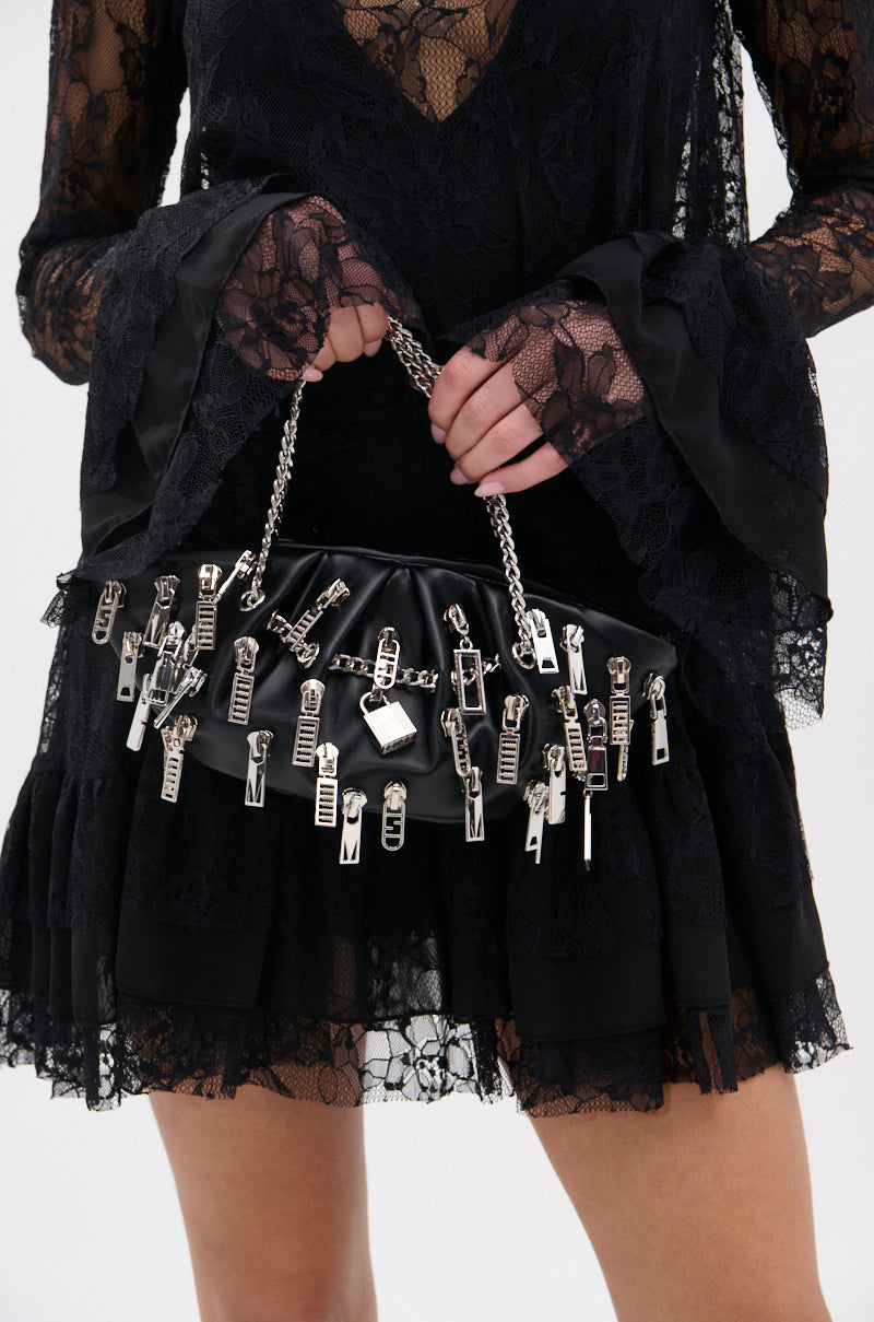 A person in a black lace dress holds the ZIP IT CHARM PURSE, a stylish purse featuring multiple silver zipper pulls and a silver chain handle. The photo highlights their hands, the purse, and the elegant lower part of the dress.