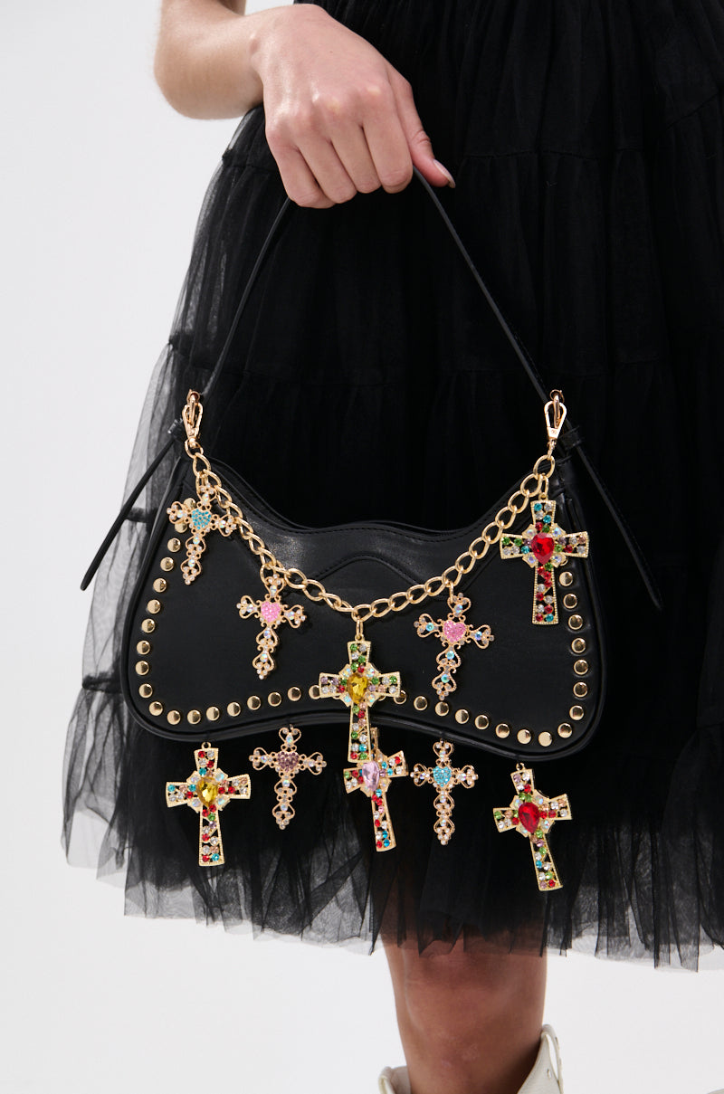 A person in a black ruffled dress holds the SO DOLCE CROSS PURSE, a black faux leather handbag featuring gold studs and a gold chain decorated with colorful jeweled cross pendants. 2