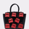 The STAY IN MY BAG PURSE has a structured silhouette in glossy faux leather, with seven small red handbags—each with a handle and gold clasp—arranged in two rows on the front of the large black bag.