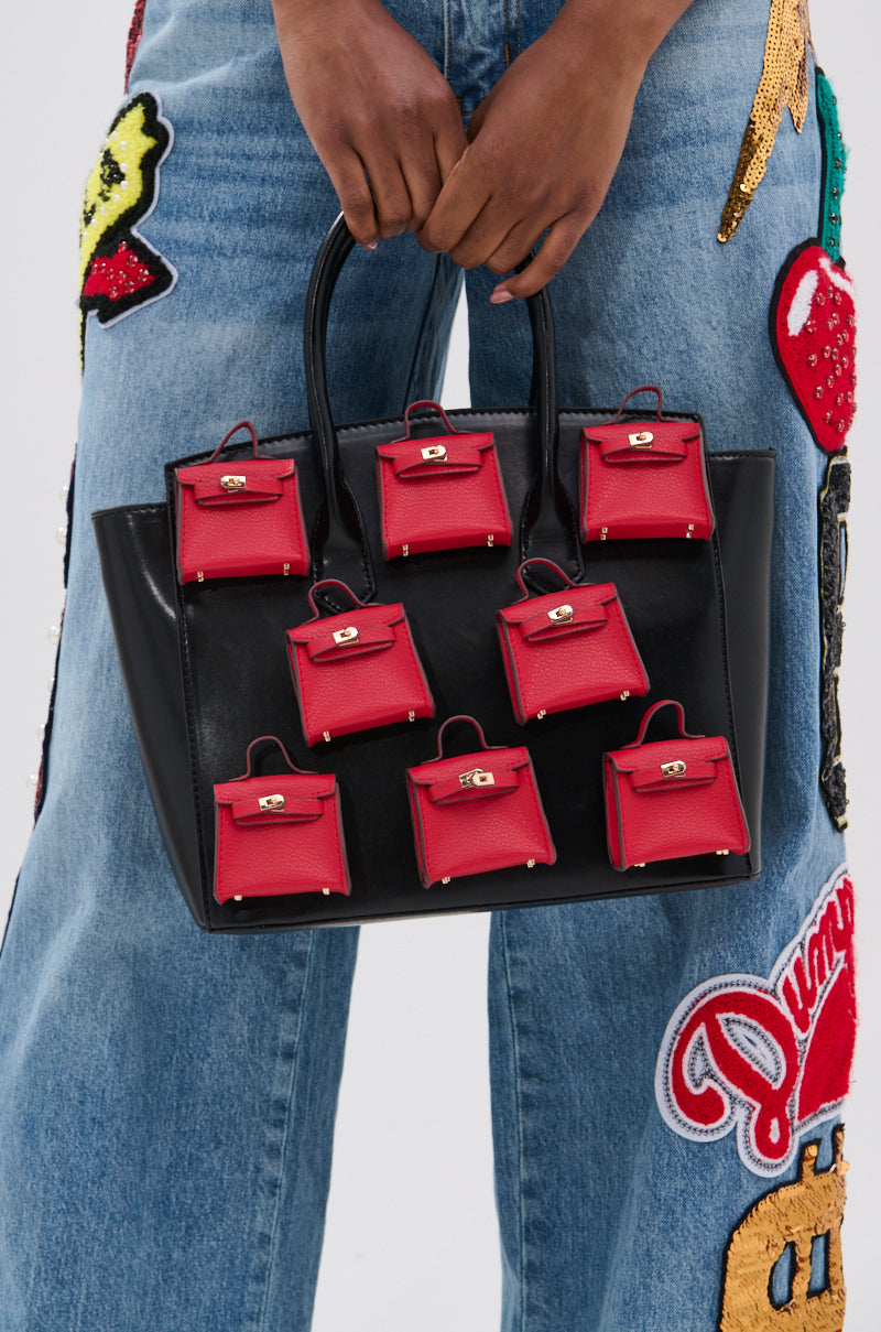 A person in embroidered blue jeans holds the STAY IN MY BAG PURSE, a black handbag adorned with several red handbag charms on its front.
