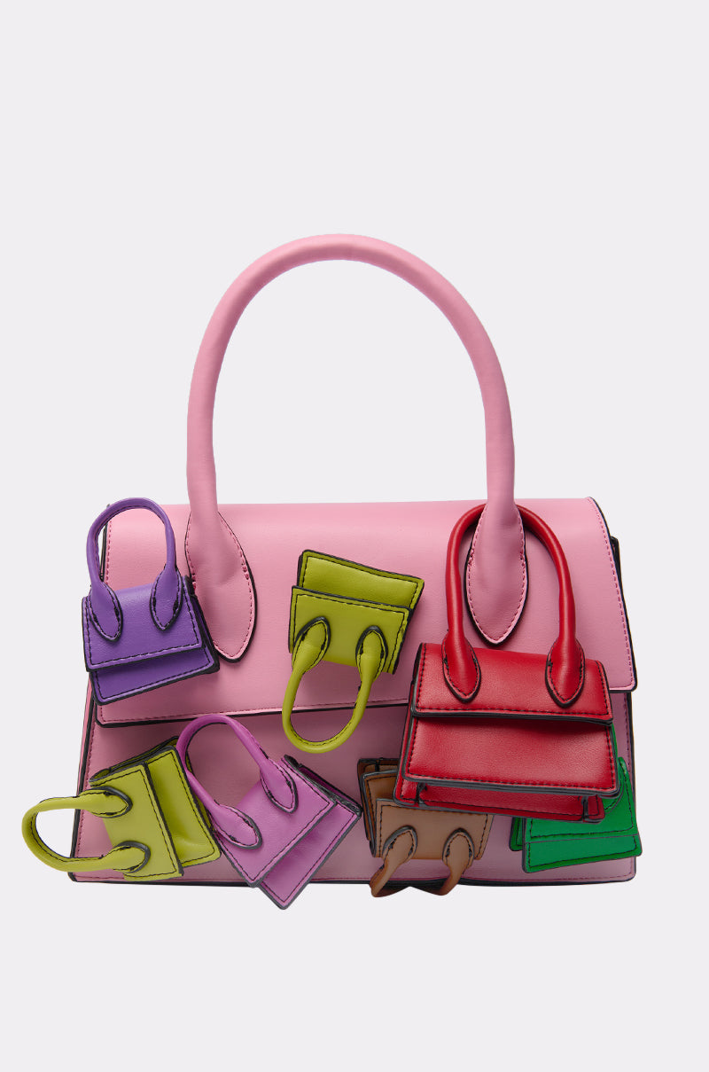 The IN MY BAGS PURSE is a large baby pink bag decorated with six colorful mini bags—purple, green, red, brown, and pink—attached to its front and handles, shown against a plain light background. 0