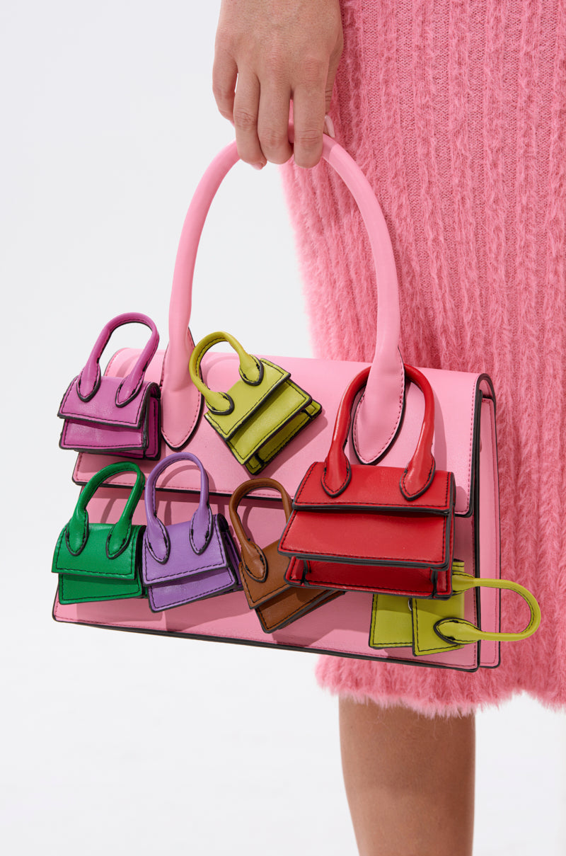 A person in a pink textured skirt holds the IN MY BAGS PURSE, a baby pink faux leather bag featuring six colorful mini purses on the front and a detachable shoulder strap for versatile styling.