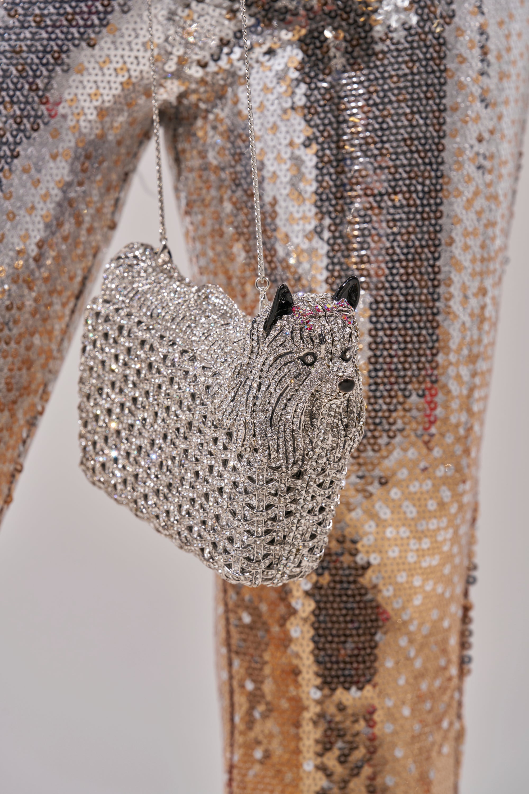 The CHARMAINE BLING PURSE, a dog-shaped accessory covered in silver rhinestones, hangs against sequined pants shimmering in gold, brown, and silver tones.