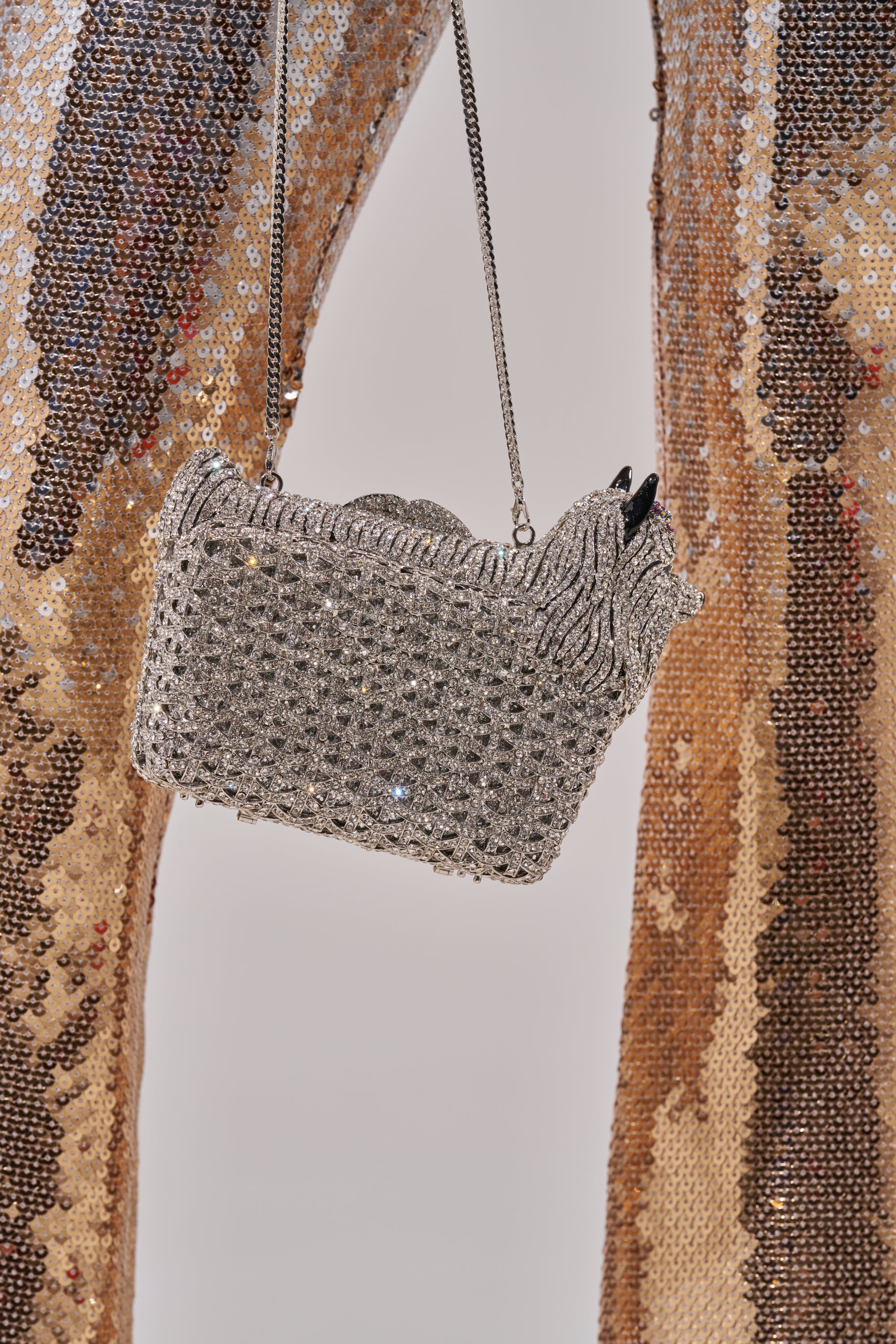A close-up of the CHARMAINE BLING PURSE, a sparkly rhinestone bag with a chain strap, held in front of gold and brown sequined pants, showcasing the shimmering textures of both the purse and the outfit.