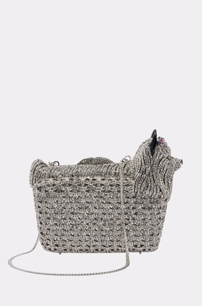 The CHARMAINE BLING PURSE is a sparkling silver, dog-shaped handbag featuring intricate rhinestone details, a chain strap, and a sculpted dog head on one end—an eye-catching statement piece against a white background.