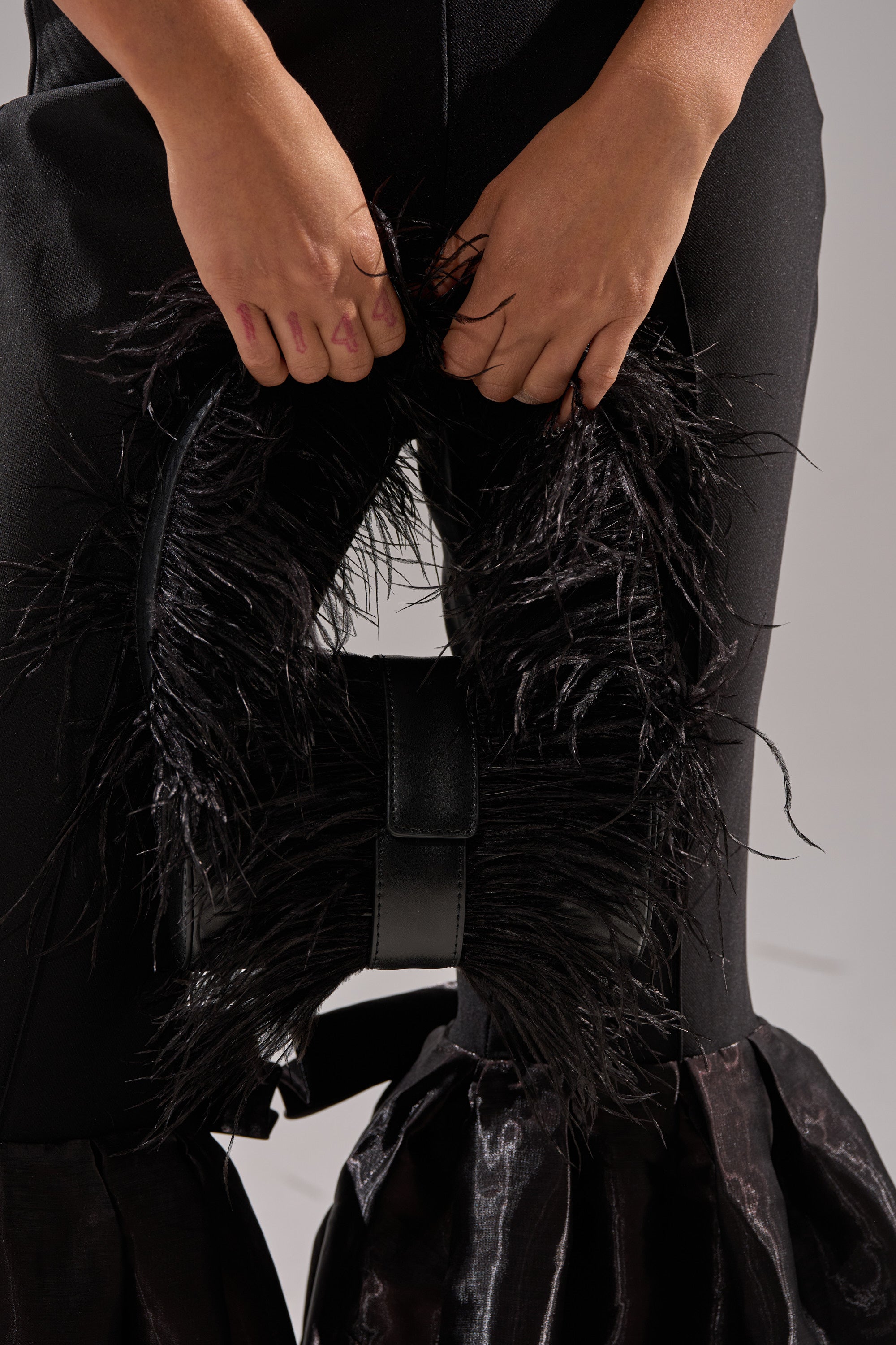 A person holds the FEATHER CRAZE PURSE—a black, furry-textured statement bag—in front of them; only their hands and lower torso, dressed in black pants with ruffled, shiny cuffs, are visible.