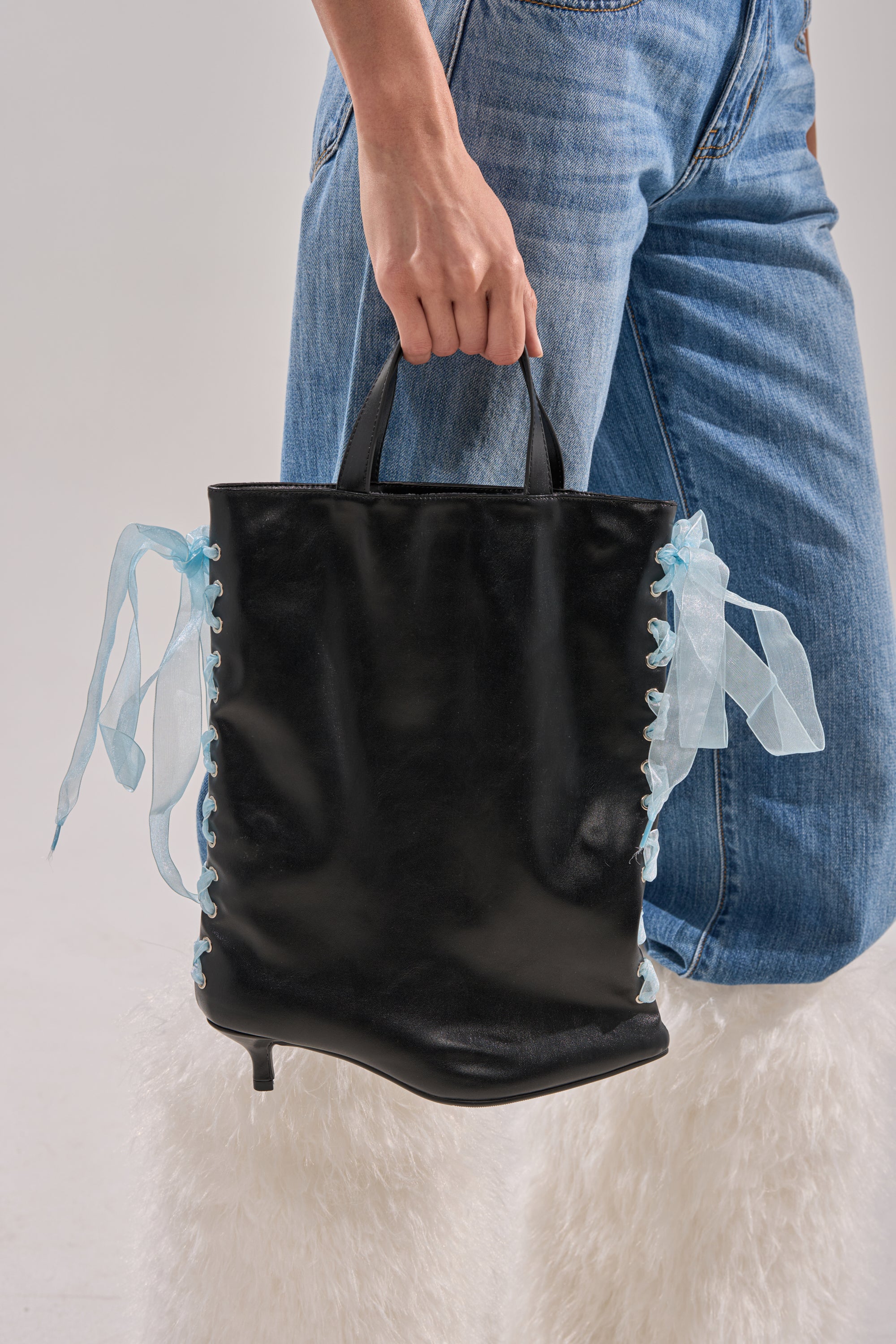 A person in light blue jeans and white furry boots holds the TIE MY SHOE PURSE, a black vegan leather bag with blue sheer ribbon lacing on the sides and small handles.