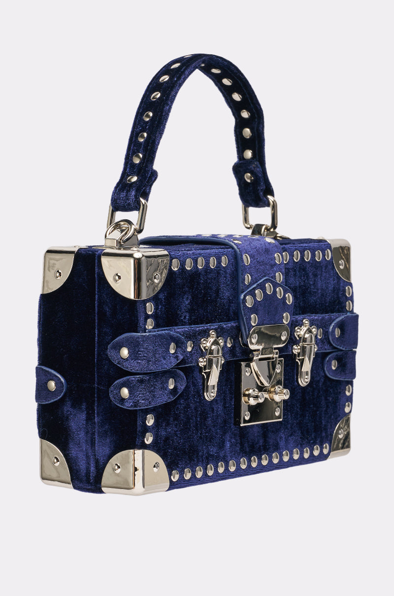 The TRUNK SHOW PURSE is a rectangular navy velvet mini trunk bag featuring silver studs, metal corner protectors, shiny buckles, and a top handle—resembling a vintage suitcase with decorative metal accents for added charm.