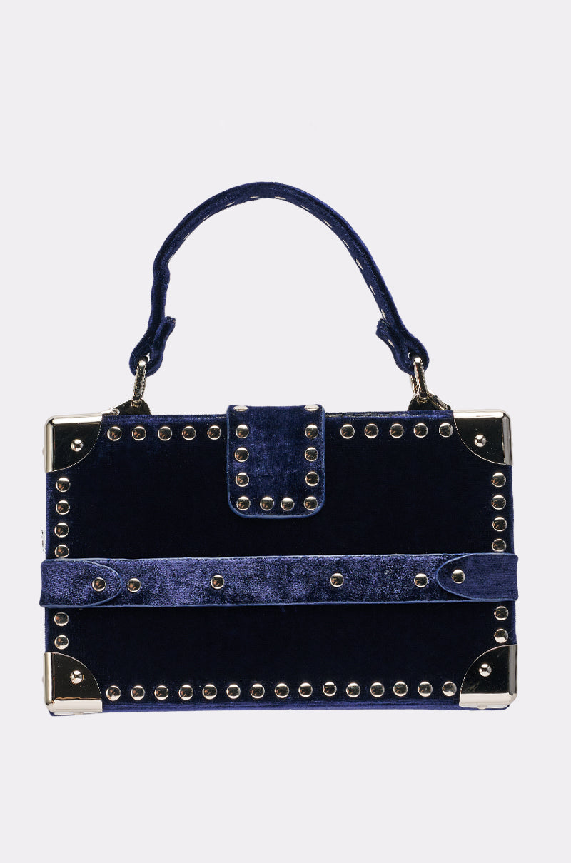 The TRUNK SHOW PURSE is a small rectangular navy blue velvet handbag with silver studs, a top handle, and metallic corner accents, inspired by vintage trunks.