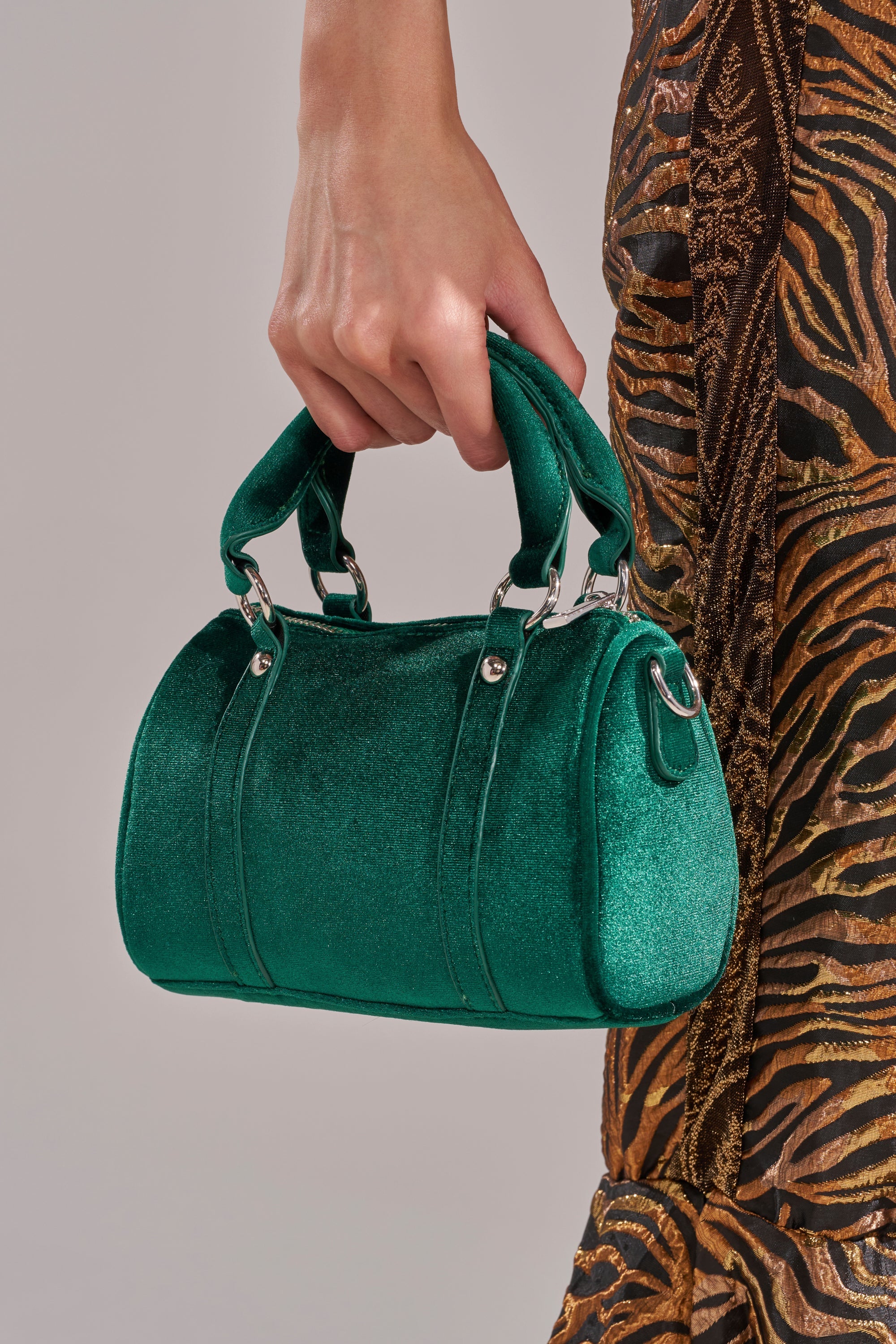 A person in a brown and black animal print outfit holds the JESSI VELVET PURSE, a small shiny green structured handbag with silver hardware and two handles, against a neutral gray background. 1