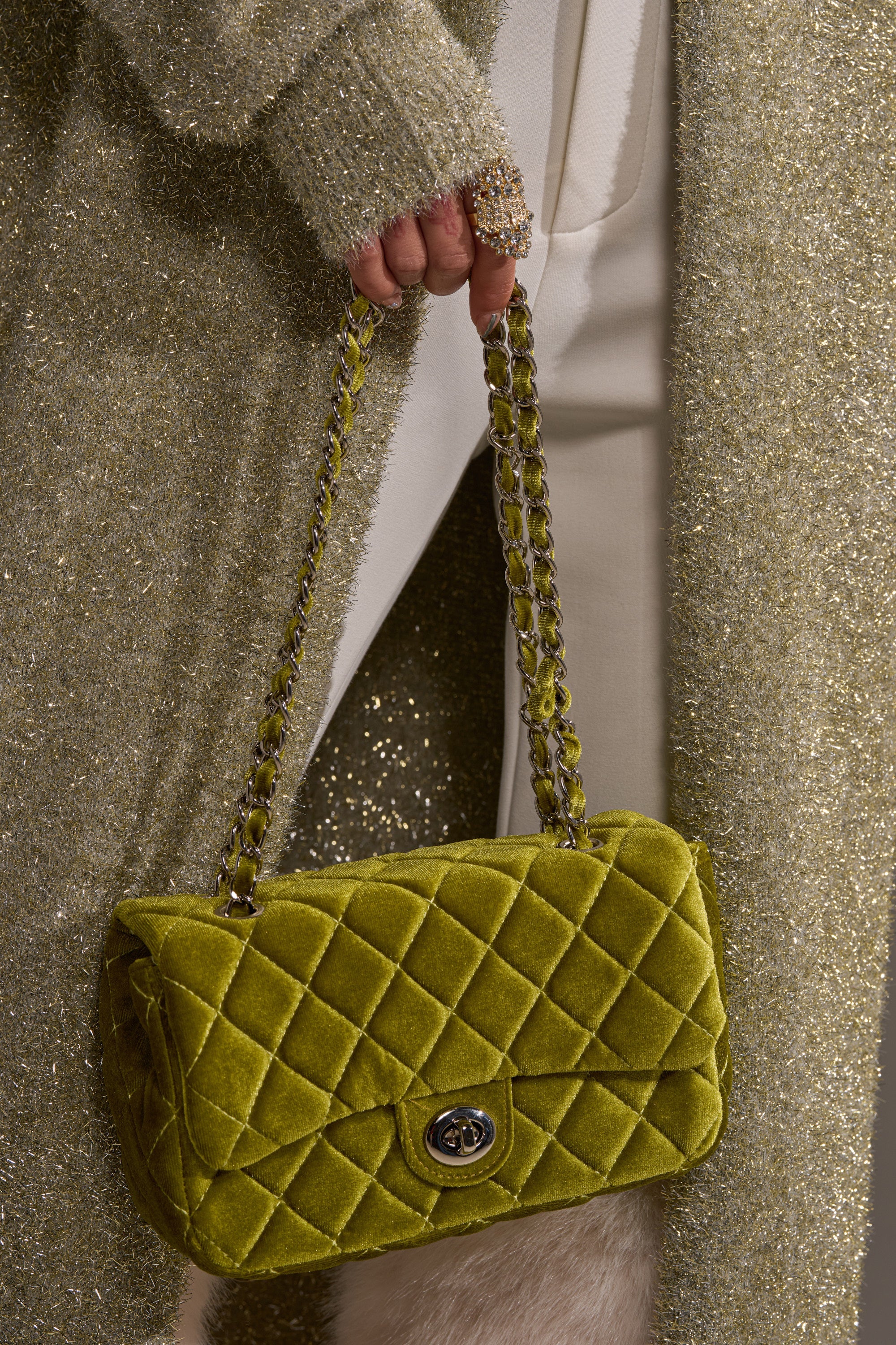 Dressed in glittery gold, a person holds the NIKKI VELVET QUILTED PURSE in olive-green, featuring a chain strap and turn-lock closure. A sparkly gold ring completes the glamorous look. 4