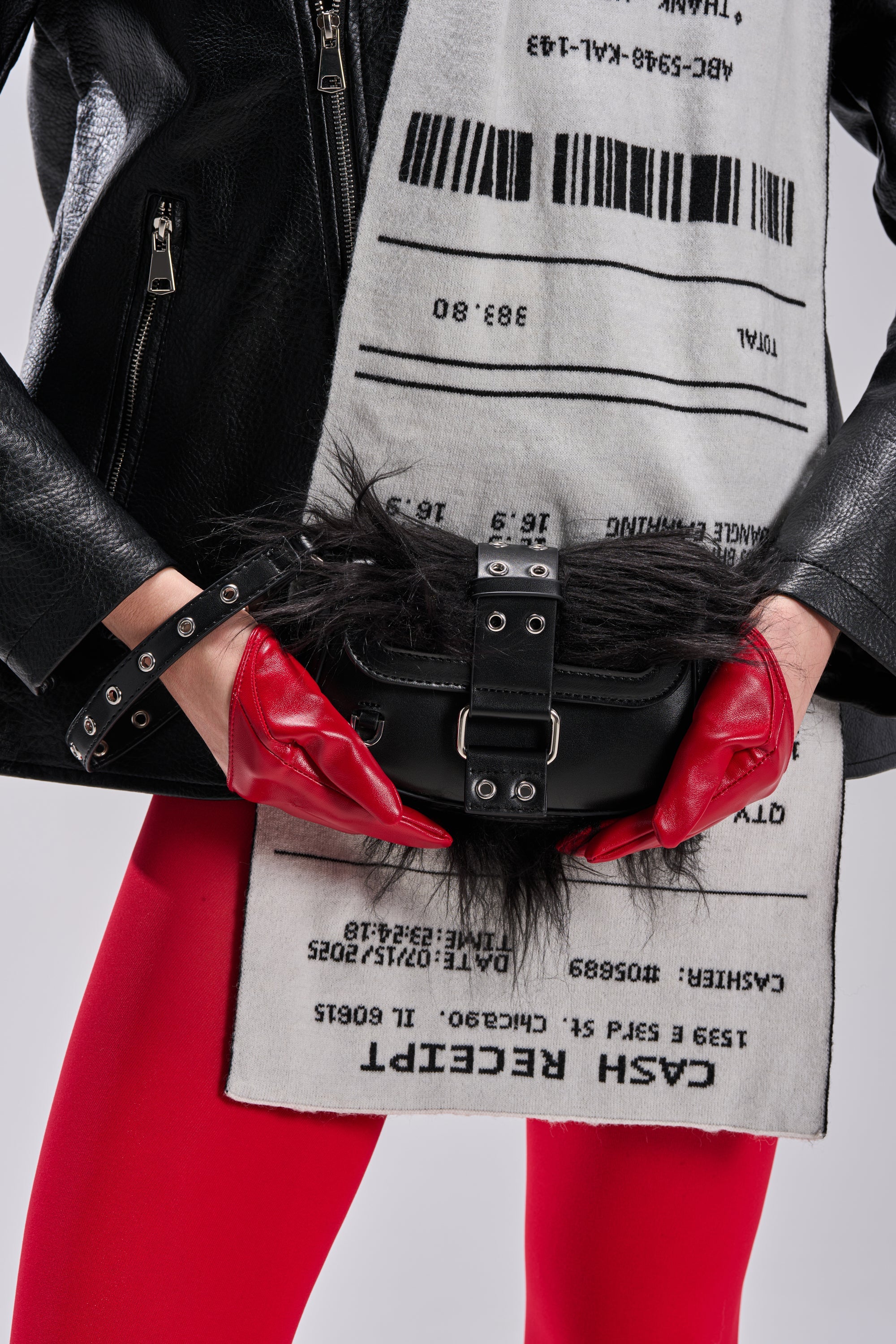 A person wears a black faux leather jacket, red gloves, and red pants, holding the ON THE TOWN WRISTLET—a black bag with fur and buckles. A white scarf featuring a barcode and receipt print completes this bold night-out look. 1