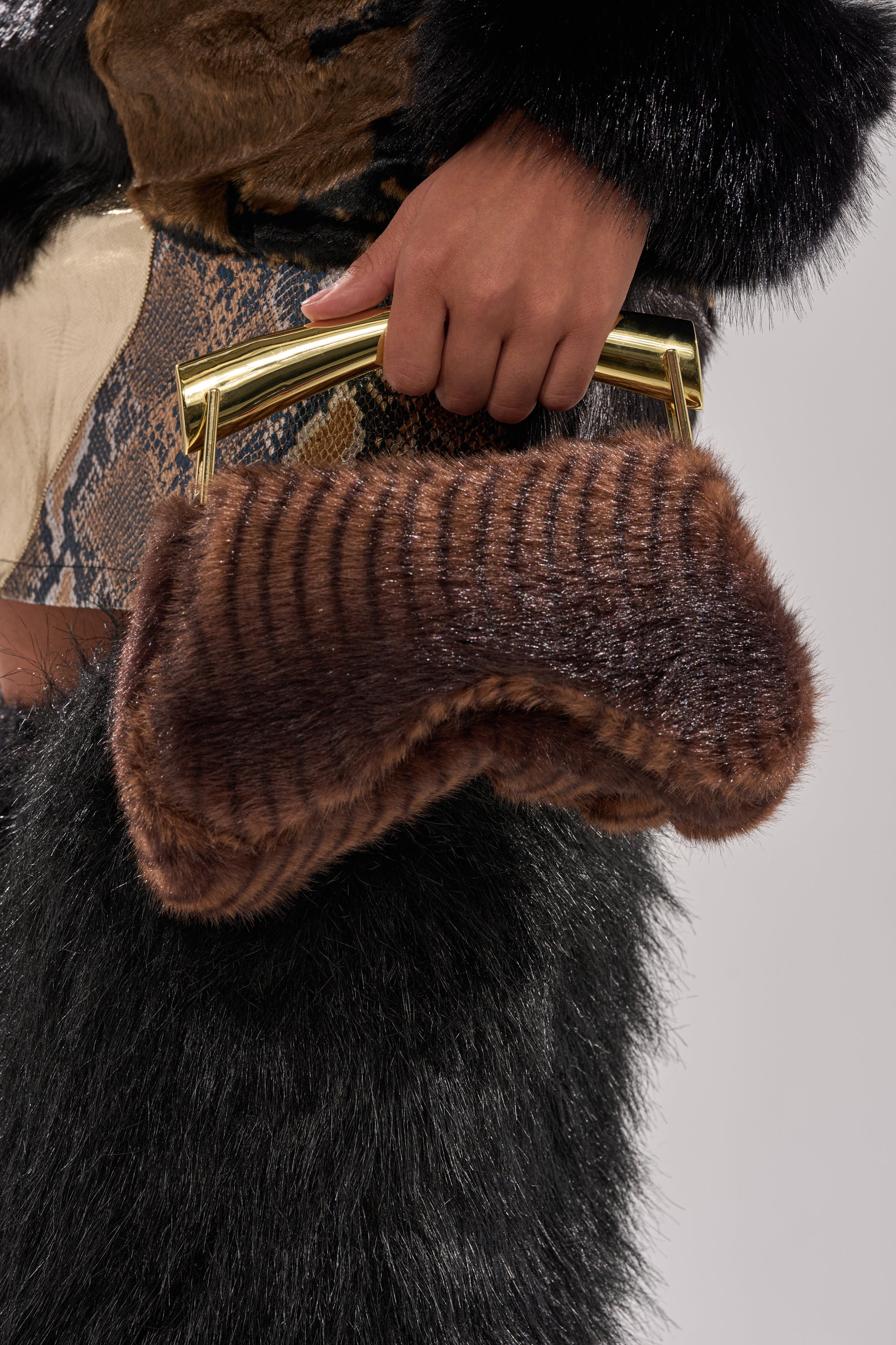 A person in a black furry coat holds the COCO FAUX FUR BAG, which is brown with a metallic gold handle, against a plain light-colored background. 1