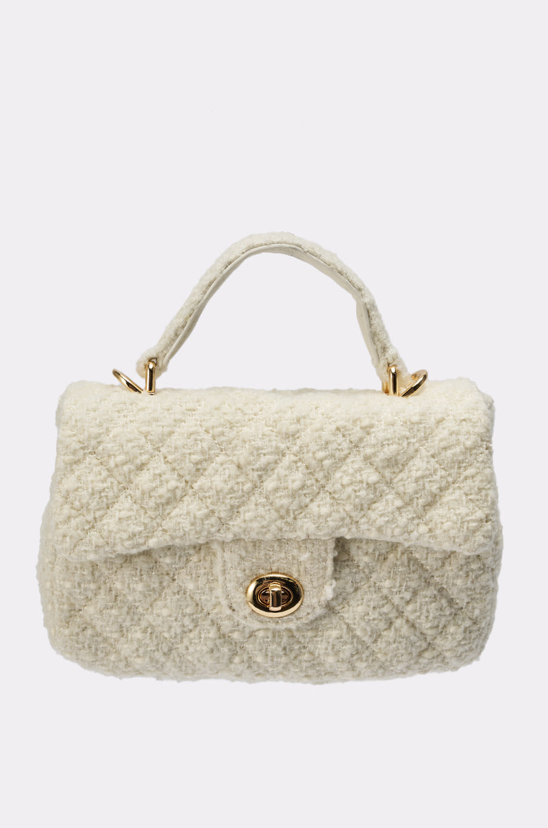 The IN MALLORCA QUILTED PURSE is a small, cream-colored mini bag with a top handle, gold-tone hardware, and a front flap twist-lock closure, displayed against a plain light background. 0