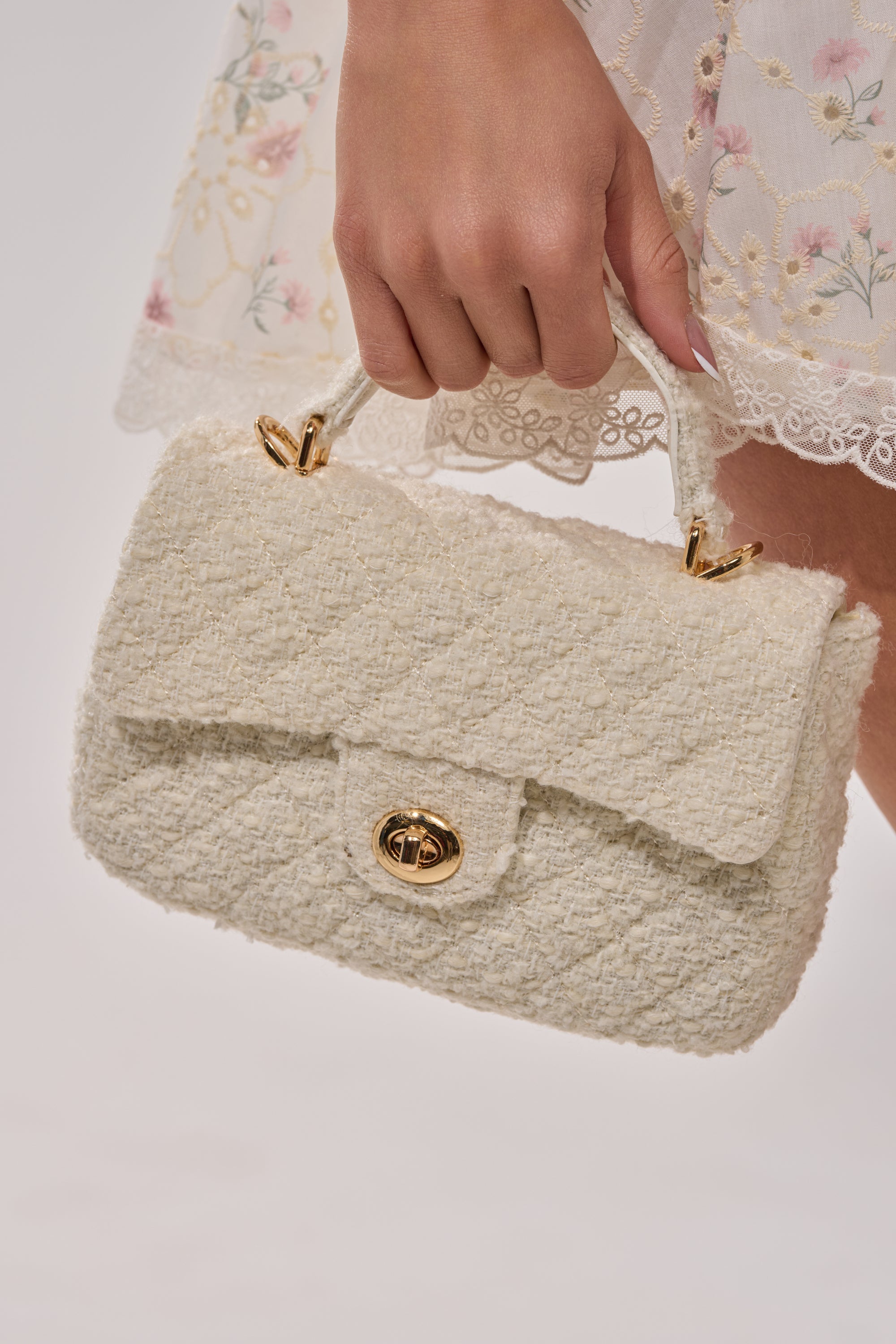 A hand holds the IN MALLORCA QUILTED PURSE with a gold clasp and handle, its ivory quilted texture standing out against a white dress embroidered with pastel floral patterns. 3