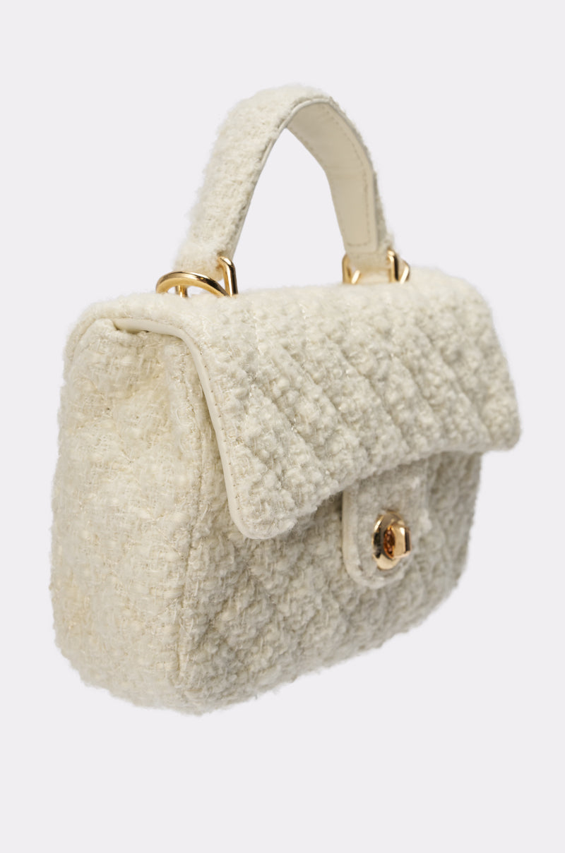 The IN MALLORCA QUILTED PURSE is a chic cream mini bag with textured quilted fabric, a short handle, gold hardware, and a round clasp on the front flap, shown against a plain light background. 2