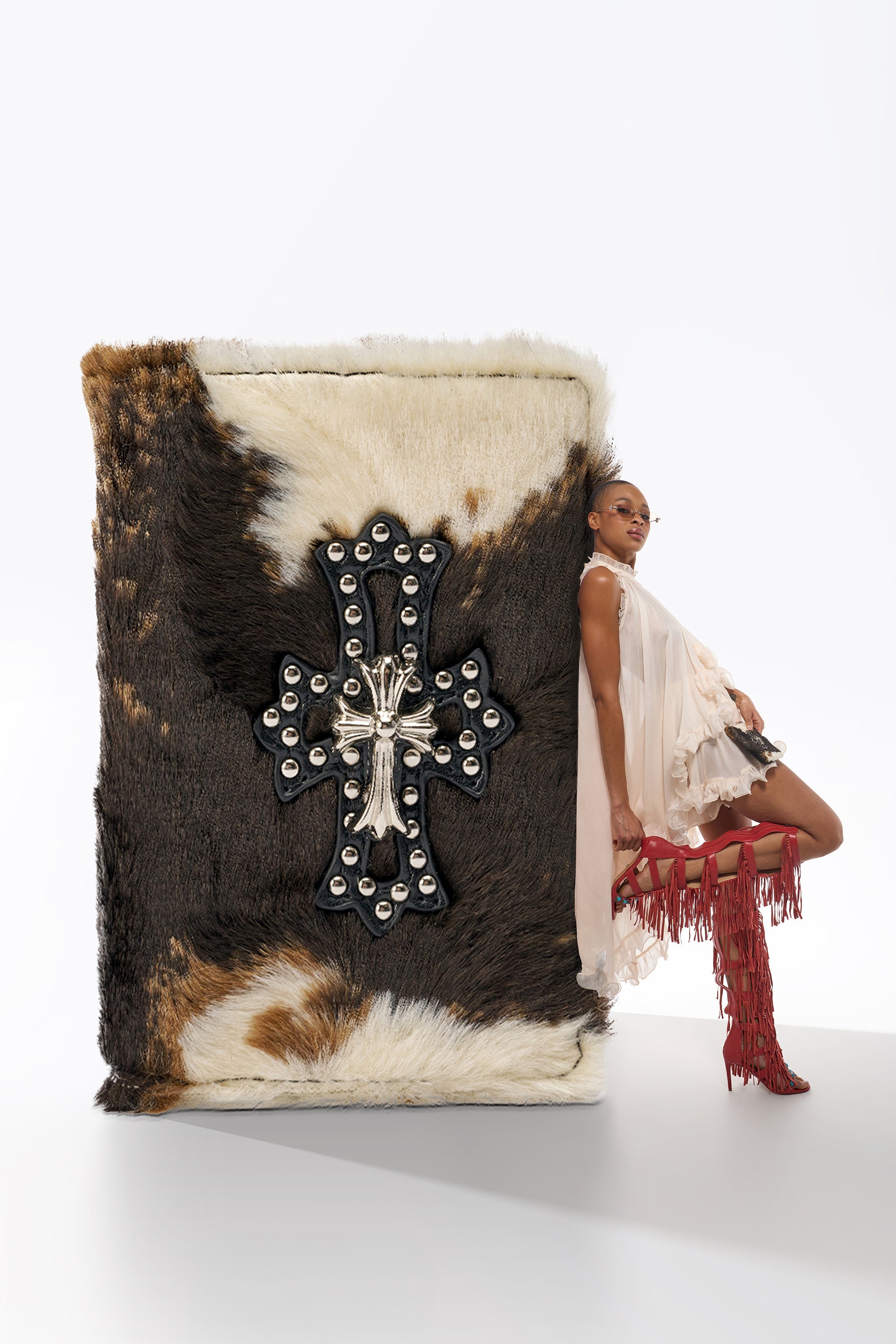 A woman in a ruffled dress and red fringe boots leans against the HIGH STANDARDS FAUX FUR PASSPORT COVER, oversized and styled as a book with a decorative cross and metallic details, set on a plain white background. 0