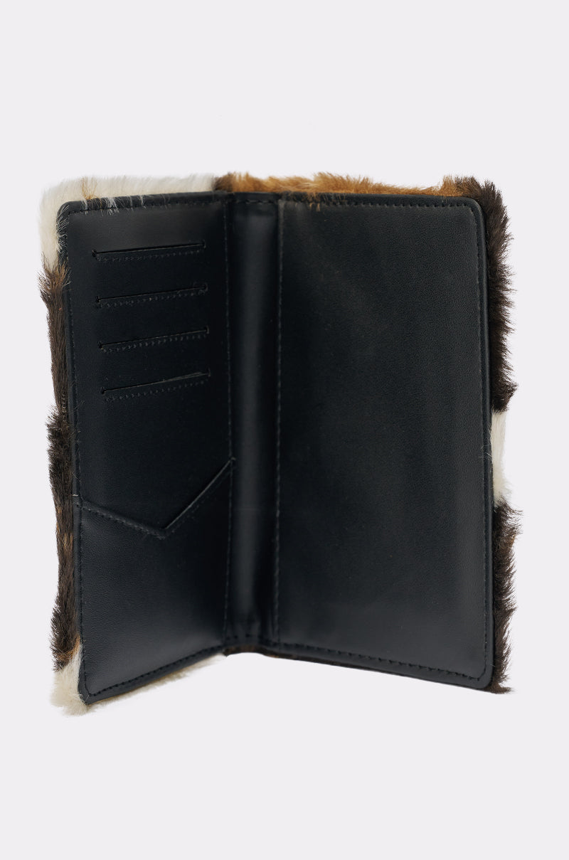 The HIGH STANDARDS FAUX FUR PASSPORT COVER features open black leather with brown and white cow print faux fur trim, card slots, and a pocket inside the left panel—perfect for keeping travel essentials organized. 2