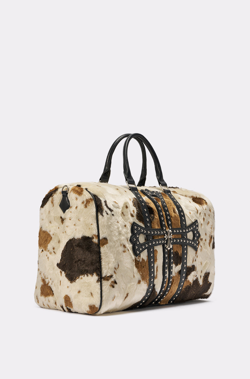 The HIGH STANDARDS FAUX FUR DUFFEL is a rectangular cow print bag featuring black handles, brown and black leather straps with metal stud accents, and an ornate cross design on the front. 2