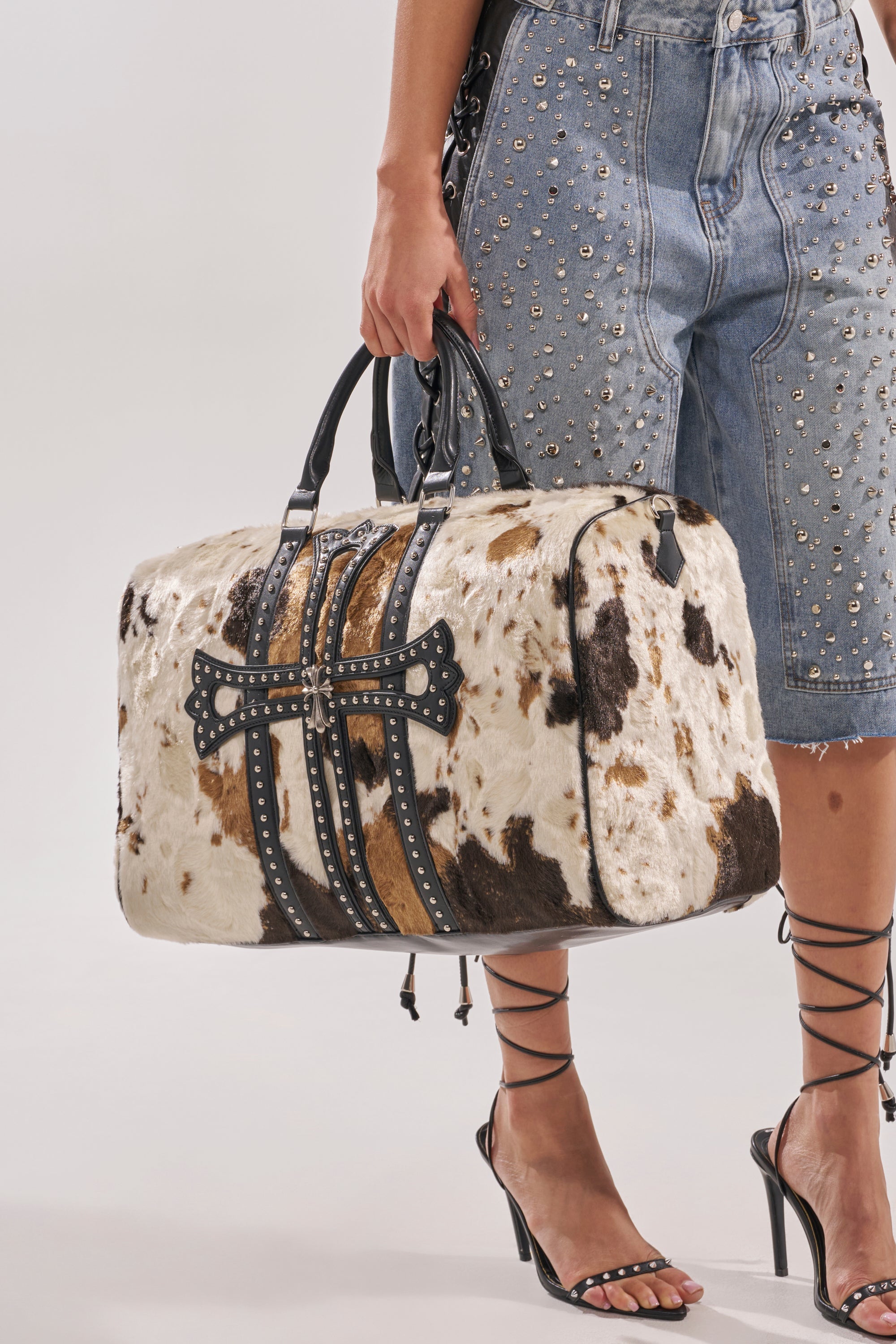 A person in studded denim shorts and strappy heels holds the HIGH STANDARDS FAUX FUR DUFFEL, a large cowhide-patterned bag with leather straps and metal studs. 6