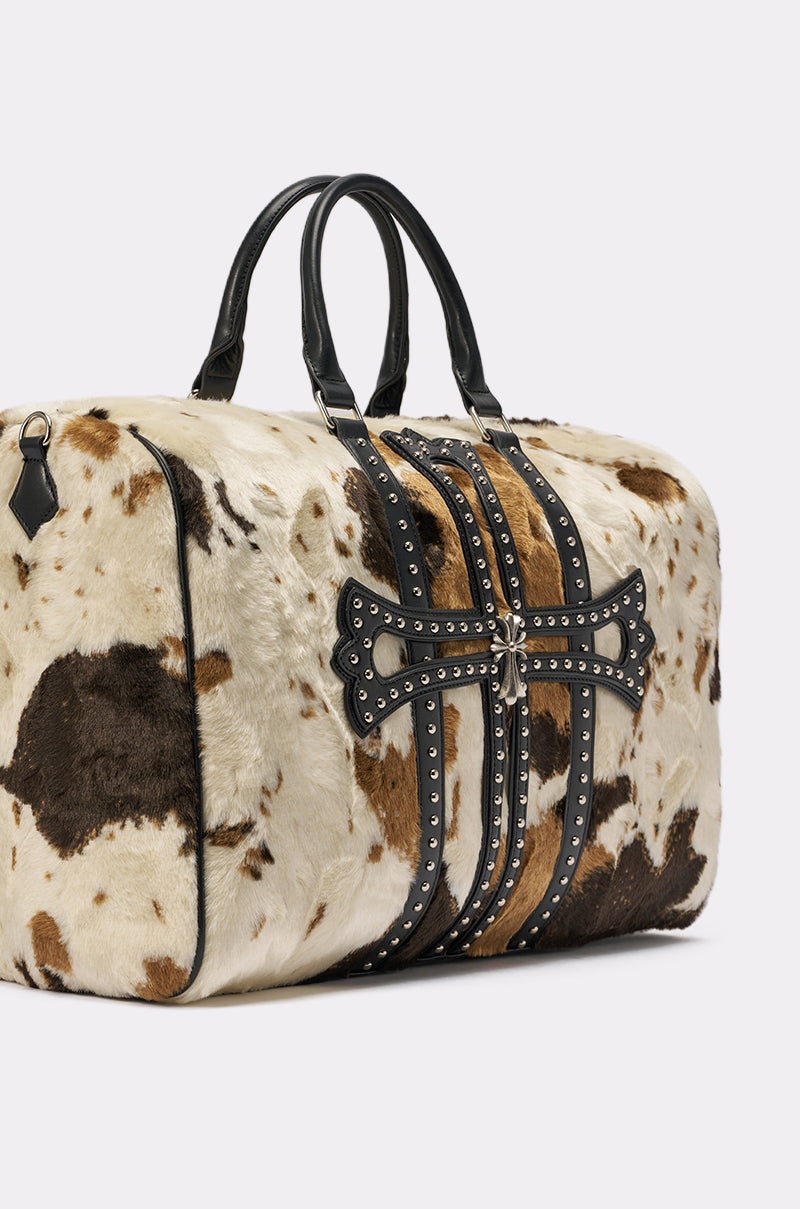 The HIGH STANDARDS FAUX FUR DUFFEL is a western-inspired cow print bag in brown, black, and white, featuring black leather handles, studded straps, and a decorative cross-shaped studded accent on the front. 4